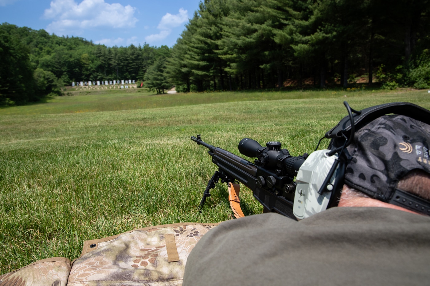 Photo taken from behind the author at an outdoor shooting range, showing the back of the author, the M1A rifle equipped with the Leupold Mark 4HD 1.5-10x scope in the foreground, and several paper or steel targets visible at varying distances downrange. The author is in a shooting position, focusing through the scope and preparing to fire. The rifle and optic are centered in the image, while range conditions and multiple target distances provide context for scope testing. The scene captures a realistic view of how the Mark 4HD is used for long-range precision shooting. This helps readers understand how the scope performs when engaging targets across different distances.