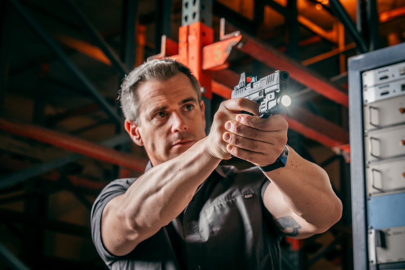 A man in casual clothing stands inside a large, industrial warehouse while holding a Hellcat Pro pistol in a two-handed grip, facing in the direction of simulated attackers. The environment is open, with visible pallets and boxes in the background, mimicking a realistic workplace or storage facility setting. The individual’s posture shows alertness, with his arms extended and body angled for defensive readiness. This scene illustrates concealed carry response training, focusing on using a compact handgun for self-defense in non-traditional or challenging environments. The photo is intended to demonstrate how situational drills improve confidence and response speed for EDC firearm users.