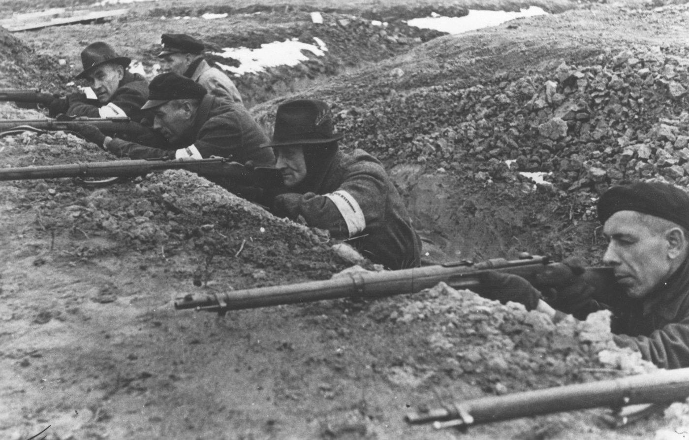 The photo shows a group of elderly Volkssturm militia members huddled in a trench, each holding a different type of bolt-action rifle, highlighting the lack of standardized weaponry in late-war German forces. The men are dressed in a mix of civilian clothes and military gear, illustrating the improvisational nature of the Volkssturm units. Their defensive posture and the rough conditions of the trench emphasize the desperate circumstances faced by Germany in 1945. The image underscores the logistical challenges encountered when arming such a diverse group of recruits. The proposed remedy, the Gustloff Volkssturmgewehr, was meant to address these equipment shortages by providing a simple, mass-produced rifle. The overall atmosphere is tense and somber, capturing the reality of last-ditch defensive efforts. The setting is bleak, with muddy earth and improvised fortifications typical of late-war trenches. This image helps contextualize the purpose behind the development of emergency weapons like the Volkssturmgewehr.