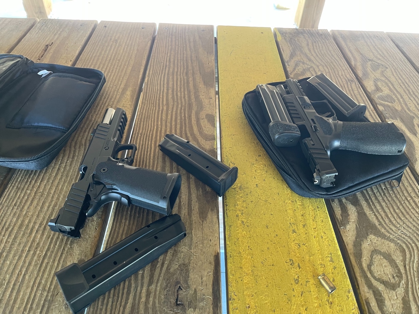 Several pistols are arranged neatly on a shooting bench along with multiple spare magazines, set up for a reloading drill at a public shooting range. The pistols appear unloaded, with magazines removed and placed in an organized row, showing a safe and controlled environment. This setup demonstrates the equipment needed for reload practice, which is a key part of many public range-friendly drills. The image highlights the importance of having extra magazines on hand to run drills like the 1-Reload-1 or Baer Solutions Standards. Visible hearing protection and shooting accessories add context to the scene, reinforcing safe firearm handling. The organized layout makes it easy for shooters to access magazines quickly during practice. This photo is ideal for articles covering reloading techniques, pistol training, and safe range habits. The overall message is preparation and safety during structured training sessions.