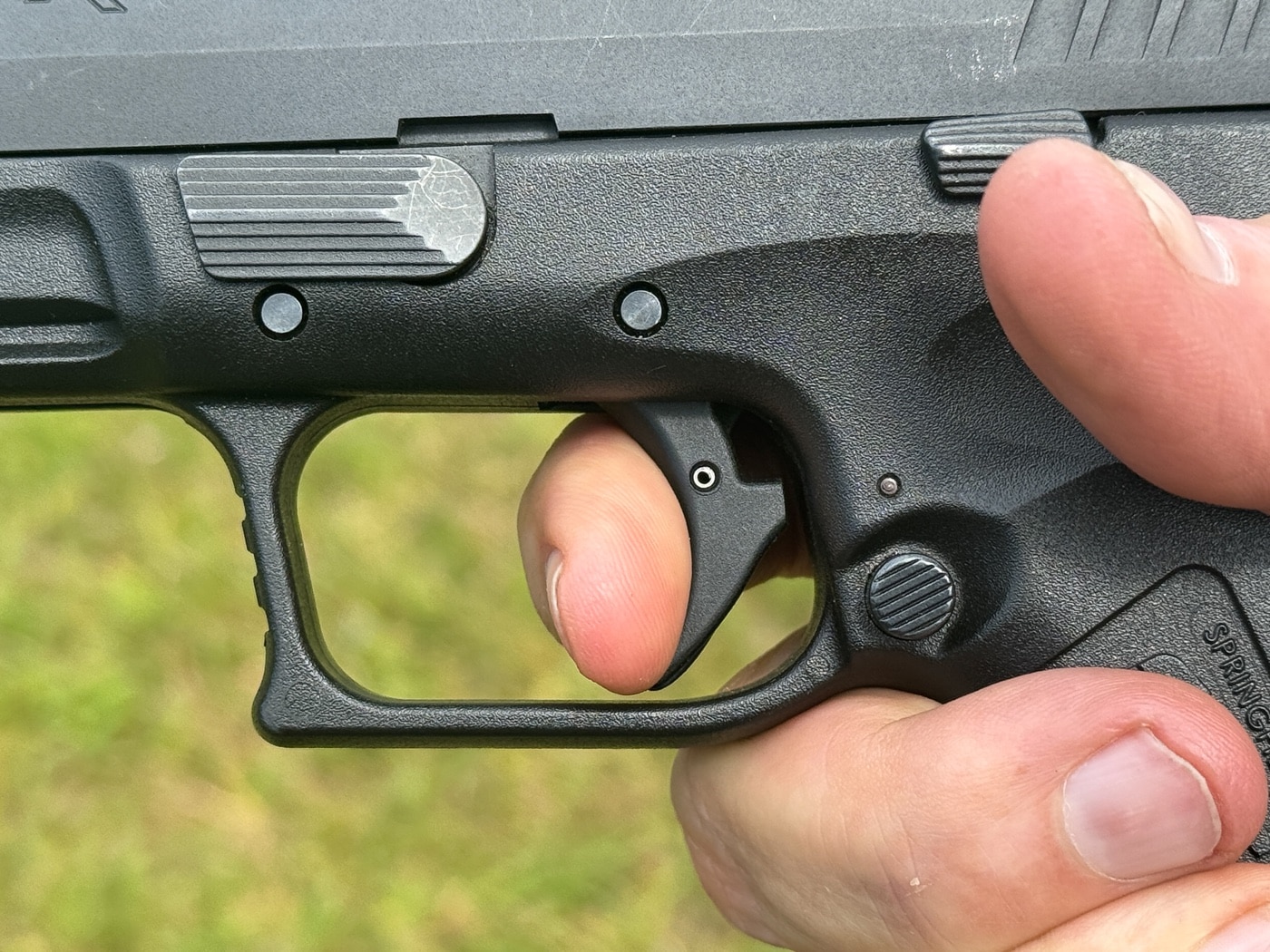 This close-up photo highlights a shooter’s trigger finger remaining in contact with the trigger as it moves just enough forward to reach the reset point. The finger does not leave the trigger face, demonstrating the riding the sear technique discussed by Massad Ayoob. The handgun is shown from the side, and you can see the subtle trigger movement required for a proper reset without letting the trigger go fully forward. The focus is on the finger position at the instant the internal reset click is felt. The background may include range details but remains uncluttered, keeping attention on the shooter’s hand and the gun’s controls. This method, as explained in the article, is used for precise control and fast follow-up shots, especially in competition or slow-fire scenarios. The image helps illustrate one of the most advanced trigger control methods along Ayoob’s bell curve of techniques.