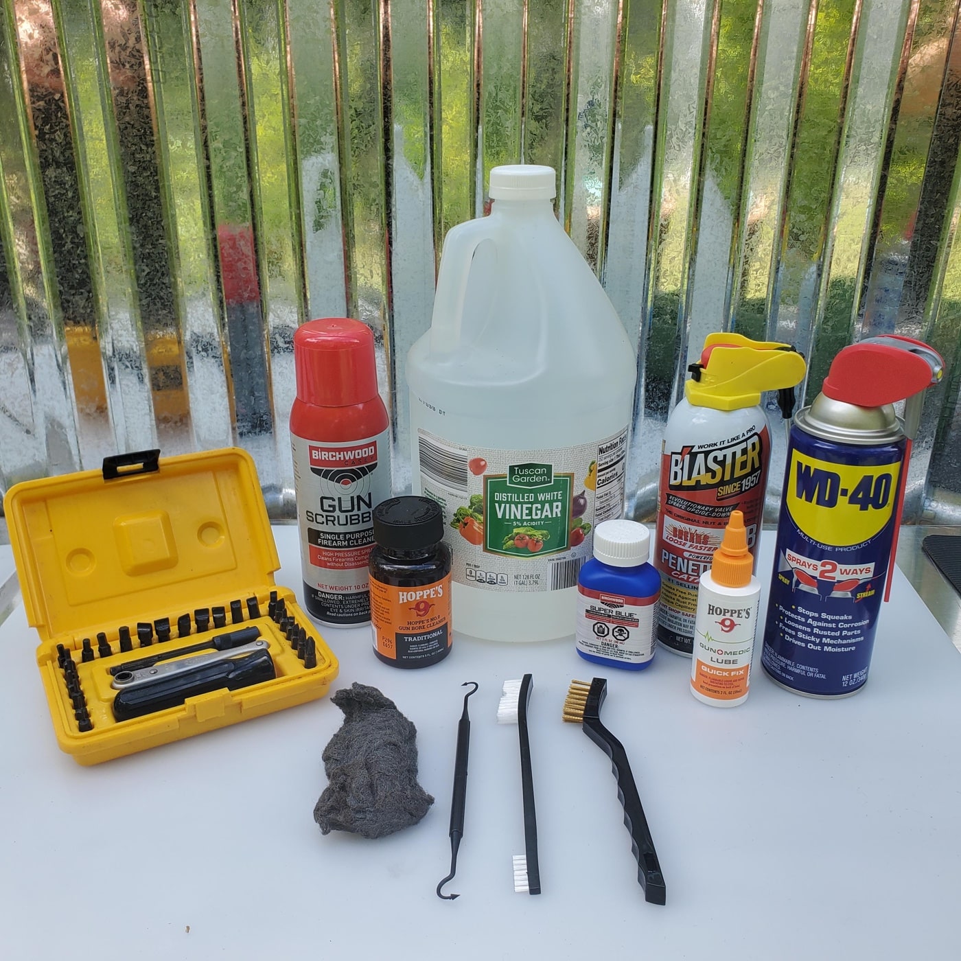 A photo displaying a group of rust removal supplies laid out on a workbench, intended for cleaning and restoring firearms. The supplies include a bottle of white vinegar, a can of penetrating oil, several pieces of fine steel wool, cleaning brushes, lint-free cloths, latex gloves, and a bottle of cold blue solution. The items are arranged to be easily visible and accessible, with some brushes and steel wool placed close together to highlight their use in scrubbing away rust. Safety glasses are included in the layout, reinforcing the importance of protection while working with chemicals. This image supports the article topic “how to remove rust from a gun” by illustrating exactly what materials are needed to do the job safely and effectively. It encourages readers to gather the correct supplies before starting any cleaning work. The setup signals that both common household and specialized products can be used for firearm maintenance.