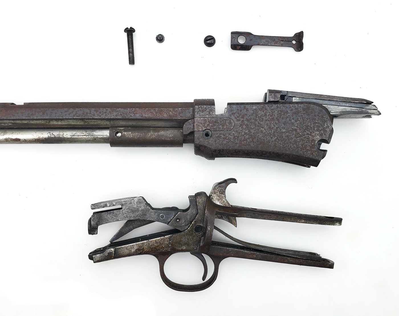 This image shows a set of disassembled gun parts spread out on a work surface. Each piece, including the receiver, barrel, screws, and smaller internal components, shows varying degrees of surface rust. The rust appears as orange and brown discoloration, particularly along edges and exposed metal surfaces. The parts are arranged in a way that makes it easy to see where corrosion is most severe and where lighter cleaning may be needed. The background is a basic workbench or table with a cloth or towel underneath to catch debris. The scene illustrates why disassembly is crucial for addressing rust — hidden areas and interior parts often need just as much attention as the outside. This photo helps readers understand the importance of inspecting all parts individually before starting the cleaning process.