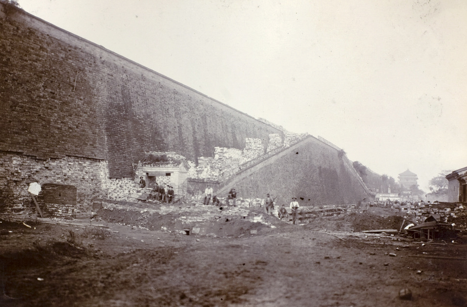 The photograph features U.S. soldiers standing at the base of the enormous Tartar Wall in Peking (Beijing) during the Boxer Rebellion, clearly dwarfed by the sheer height and thickness of the ancient structure. The wall, built of massive stone or brick, rises far above the soldier’s head — its surface marked by centuries of weathering, with possible crenellations or parapets visible at the top. The ground at the foot of the wall may be bare earth or paved stone, and the soldier is in full field uniform, providing a sense of scale against the monumental city defenses. The image captures the intimidating nature of the wall, which posed significant challenges to both foreign troops and Chinese attackers during the siege. Background details could include other soldiers, equipment, or parts of the city, but the focus remains on the dramatic contrast between human and architecture. This photo serves as a visual explanation of why the Tartar Wall was a strategic focal point in the Boxer Rebellion.