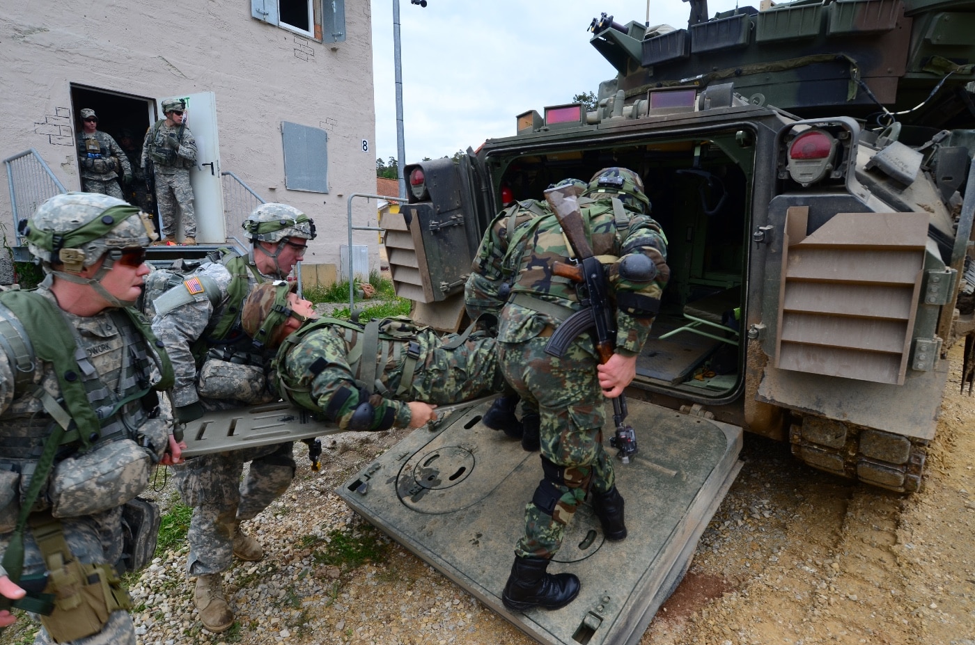 In this image, Albanian, Armenian, and U.S. Army soldiers are shown loading a simulated casualty into the rear compartment of an M2 Bradley infantry fighting vehicle during exercise Combined Resolve II at Hohenfels, Germany, in 2014. The soldiers, wearing their respective national uniforms and combat gear, are working together to lift a training mannequin on a stretcher. The open rear ramp of the Bradley is visible, providing access for medical evacuation practice. The setting is an outdoor training area, with gravel and vegetation around the vehicle. This scenario is part of a multinational exercise aimed at improving cooperation and response during medical emergencies on the battlefield. The image highlights the M2 Bradley’s capacity to serve as both a combat platform and casualty evacuation vehicle. Teamwork and quick action are key themes captured in this drill.