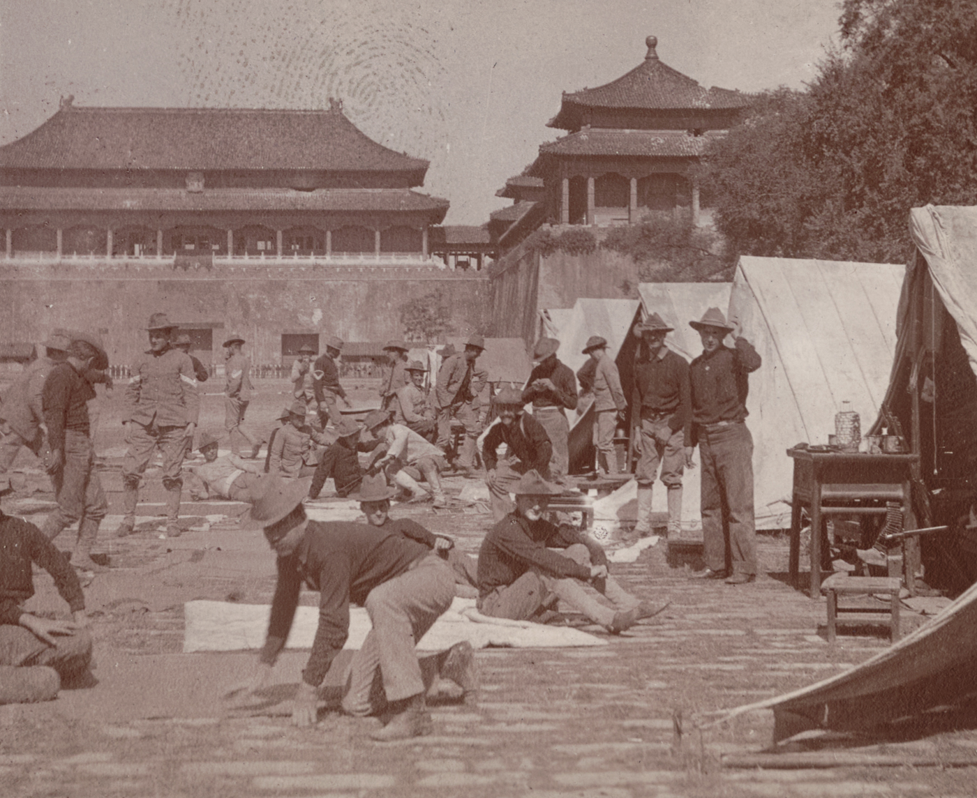 The photograph shows soldiers of the U.S. 9th Infantry Regiment camped in a spacious courtyard inside the Forbidden City in Beijing, China, shortly after the relief of the city in 1900. The men are dressed in U.S. Army field uniforms and are seen pitching tents, stacking supplies, and preparing meals amid the grand stone and tile architecture of the imperial palace. Ornate roofs, decorative pillars, and traditional Chinese gateways are visible in the background, contrasting sharply with the temporary military encampment. Soldiers sit or stand near their tents, some resting and others maintaining weapons or cooking over small fires. The historic setting underscores the dramatic change from imperial exclusivity to foreign military occupation. The photo vividly illustrates the international aftermath of the Boxer Rebellion and the U.S. Army’s unique role within China’s most famous landmark.