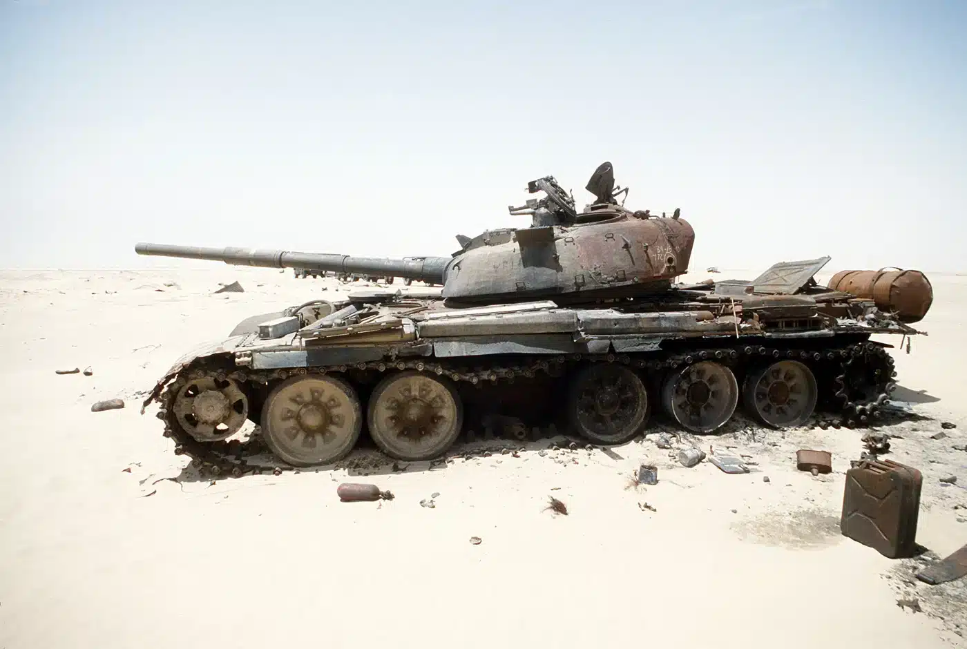 The image shows a destroyed Iraqi T-72 main battle tank near Ali Al Salem Air Base, photographed during Operation Desert Storm in 1991. The tank’s hull and turret are heavily damaged, with scorch marks and debris scattered around the wreckage. The vehicle is situated in a barren, desert environment, typical of the Kuwaiti and Iraqi battlefields during the Gulf War. Parts of the tank’s tracks and road wheels are either missing or misaligned, and the main gun appears disabled. In the background, the sandy terrain stretches into the distance under a clear or hazy sky. The photo illustrates the effectiveness of coalition anti-armor tactics, with M2 Bradleys and other vehicles credited for many such kills. This image is a stark reminder of the destructive nature of armored warfare during Desert Storm.