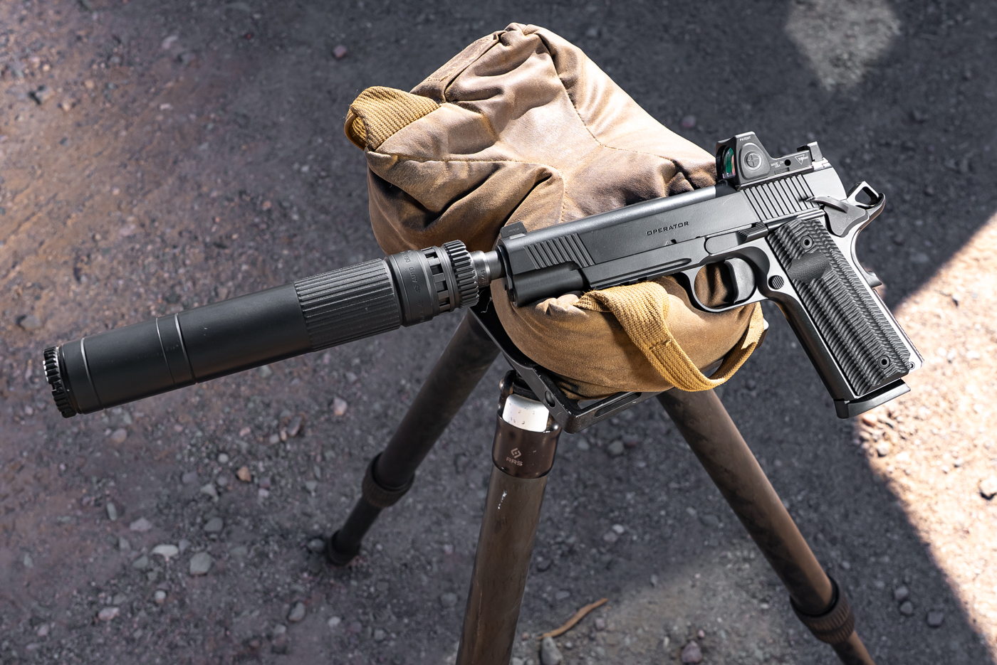 This image displays a fully equipped Springfield Armory 1911 Operator AOS Threaded .45 ACP on a range or table surface. The pistol has a Dead Air Mojave suppressor securely mounted to its threaded barrel, making the setup longer and giving it a distinctive appearance. A red dot sight is also visible, mounted at the rear of the slide using the AOS plate system. The pistol’s Cerakote finish, front and rear cocking serrations, and ambidextrous controls are clearly shown. The suppressor and optic setup illustrate how the Operator is configured for both modern optics and sound suppression. The overall presentation is professional, focusing on the functional upgrades. No hands or other objects are present in the image.