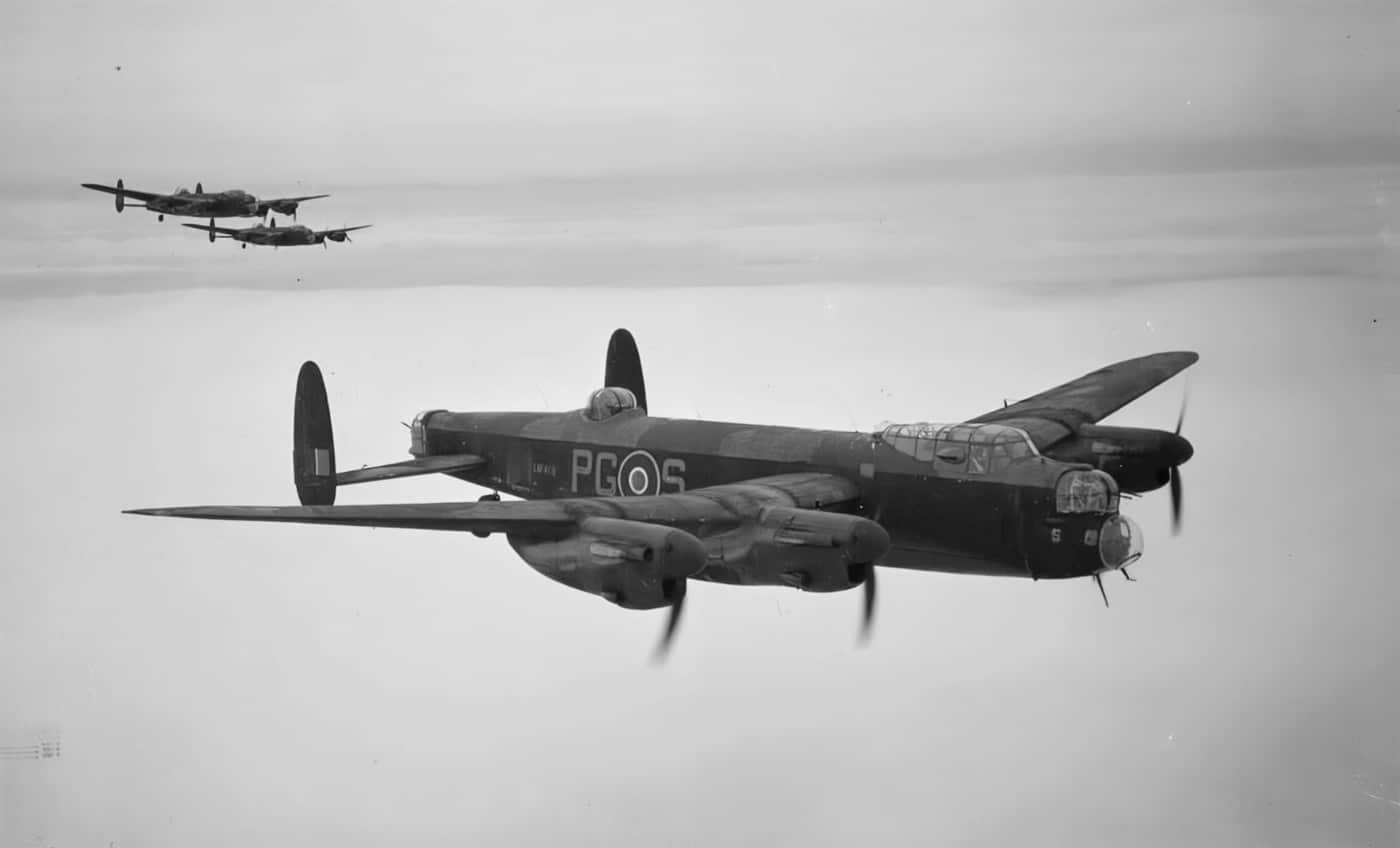 The image features three Avro Lancaster B Mark III heavy bombers flying in tight formation over the English countryside. Each aircraft is clearly visible with its four engines, straight wings, and twin tailfins. The countryside below shows typical British rural scenery with fields, hedgerows, and scattered farm buildings. The Lancasters are photographed from above or at an angle that allows viewers to see both the aircraft and the landscape below. This scene captures the operational reality of Bomber Command missions, where multiple bombers would depart together for raids on enemy targets. The photo emphasizes the teamwork, scale, and technical uniformity of the Lancaster fleet. The weather appears clear, offering good visibility of both the planes and the ground beneath them. The image serves as a strong visual for articles about RAF operations, British aviation, or the history of the Lancaster B Mark III during WWII.
