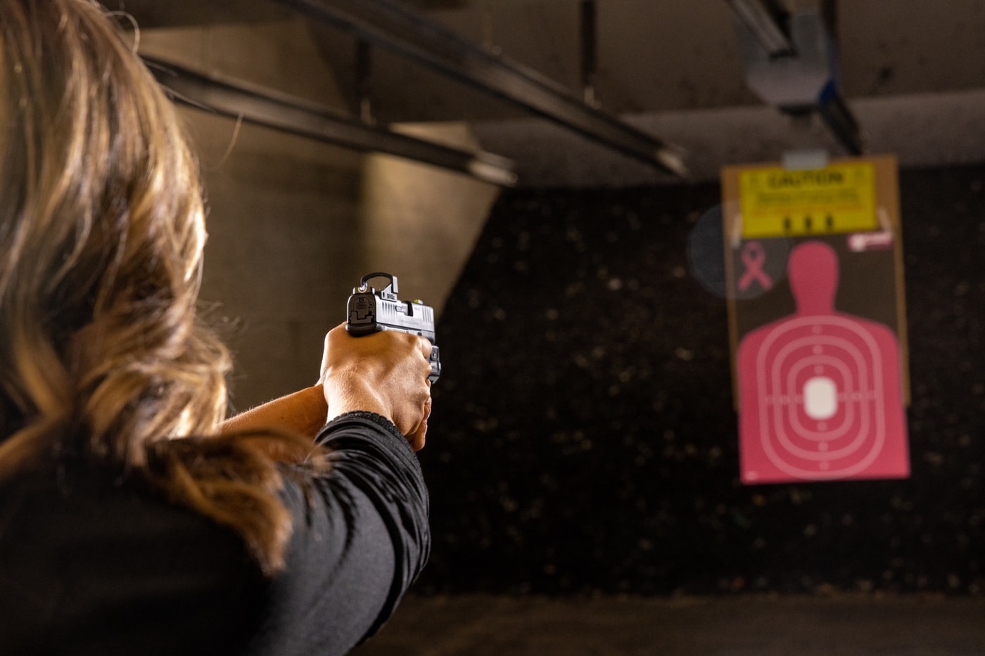 A woman stands at an indoor public shooting range, aiming a Springfield Armory Hellcat Pro pistol downrange. She maintains a firm two-handed grip and uses proper stance, focusing intently on her target. The scene shows common range safety practices like eye and ear protection, a marked firing lane, and clear line of sight to paper targets. This image is suitable for articles about pistol training at public shooting ranges, firearms safety, or self-defense shooting drills. The Hellcat Pro is a compact, popular handgun for concealed carry and practice. The woman’s posture and focus emphasize building fundamental skills like trigger control, sight alignment, and follow-through. The environment reinforces the importance of structured, safe training for all skill levels.