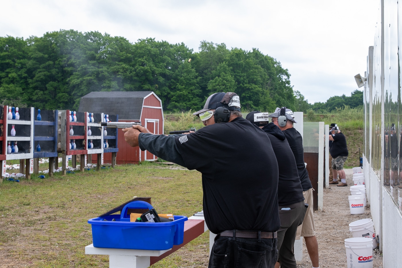 The photo captures the preliminary stage of the Springfield Armory Shootoff, where three competitors are lined up side by side at the firing line. Each shooter is in the act of aiming or firing at their respective pin tables, working to clear the pins as quickly as possible. The shooters are spaced evenly, each with their own set of bowling pin targets set up on tables downrange. Timekeepers or match officials stand just behind the competitors, monitoring progress and recording times. The setting is an outdoor range, with visible details such as range markers, safety barriers, and possibly some audience members or event signage in the background. All participants are wearing hearing and eye protection. This image highlights the unique three-person shootoff format used in the preliminaries, emphasizing the competitive and fast-paced nature of the early rounds.