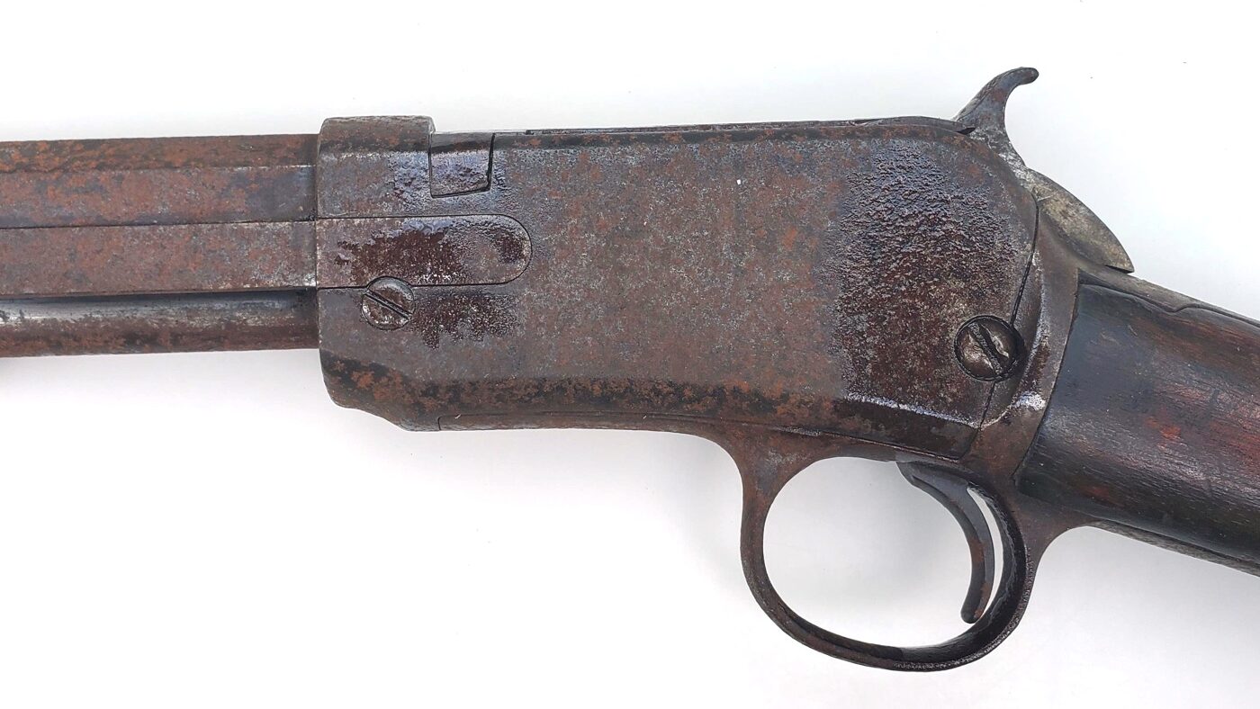 The image shows a close-up of a hand applying penetrant oil from a small can directly onto the screws of a firearm receiver before disassembly. The nozzle is aimed carefully at the screw heads, and a drop of oil is visible on the metal surface, indicating that the penetrant is being used to loosen rust or corrosion. The firearm is resting securely on a padded work surface, and the lighting highlights the rusty screws and affected areas. This photo illustrates a key step in “how to remove rust from a gun” — preparing stuck or rusted screws so they can be removed without stripping or breaking. Using a penetrant at this stage makes the rest of the cleaning process much easier and protects the firearm from unnecessary damage. The image is intended to educate readers on proper prep for gun maintenance, emphasizing careful technique and attention to detail.