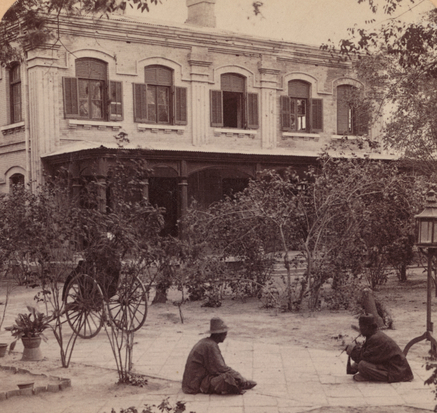 The photograph shows the western facade of the United States Legation building in Peking (Beijing) after the Boxer Rebellion, with numerous bullet holes and impact marks dotting the brick or stone exterior. The damage is concentrated around windows, doors, and architectural details, clearly revealing where gunfire and possibly shrapnel struck during the siege. Some broken windowpanes or boarded-up sections indicate hasty repairs made after the fighting. Debris may be scattered along the base of the wall, and the area is quiet, hinting at the building’s survival after intense attacks. The American flag or other legation symbols could be visible, signaling the building’s diplomatic role. This image captures the physical toll of the 55-day siege on the Legation Quarter and serves as a stark reminder of the dangers faced by the defenders. The battered wall stands as evidence of both the violence of the Boxer uprising and the resilience of those inside.
