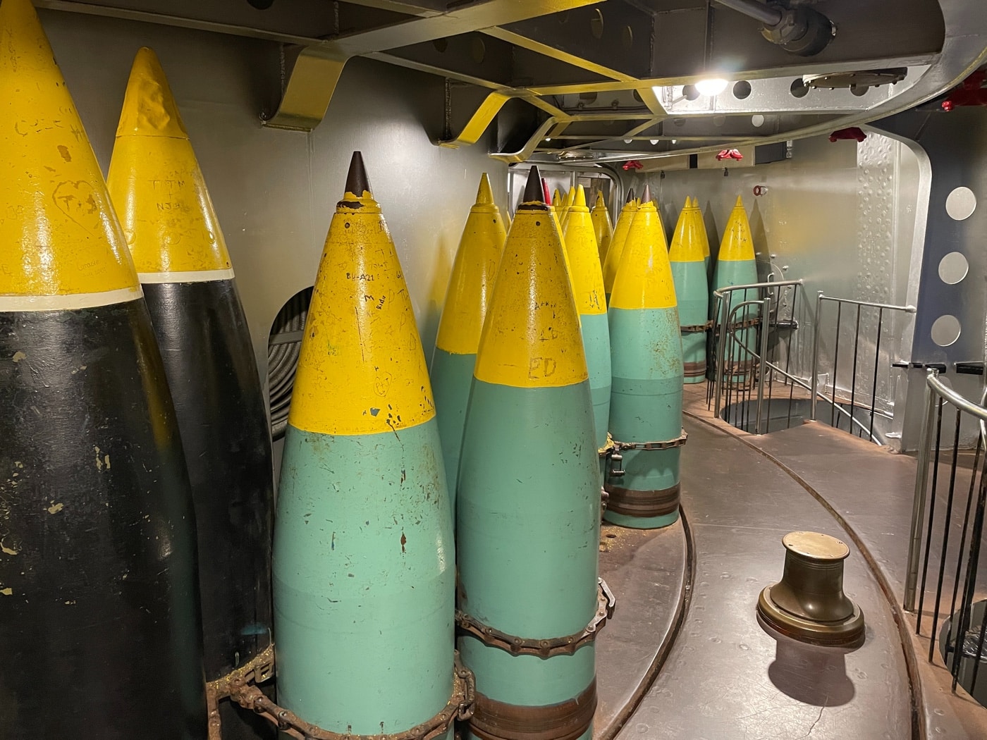 Color or black and white photo of a 2,600-pound 16-inch naval projectile, like those fired from the USS Alabama’s main battery. The shell is displayed upright, likely outdoors at the battleship memorial, with informational signage or a plaque nearby. The projectile’s steel casing is painted, possibly marked with identification or technical data. The background includes parts of the battleship’s hull or deck, helping provide context for the shell’s enormous size. Visitors or staff may be present to give a sense of scale, showing just how large each round really is. This image demonstrates the kind of ammunition used to deliver high-velocity, long-range firepower in World War II. The photo helps connect technical details with real-world hardware.