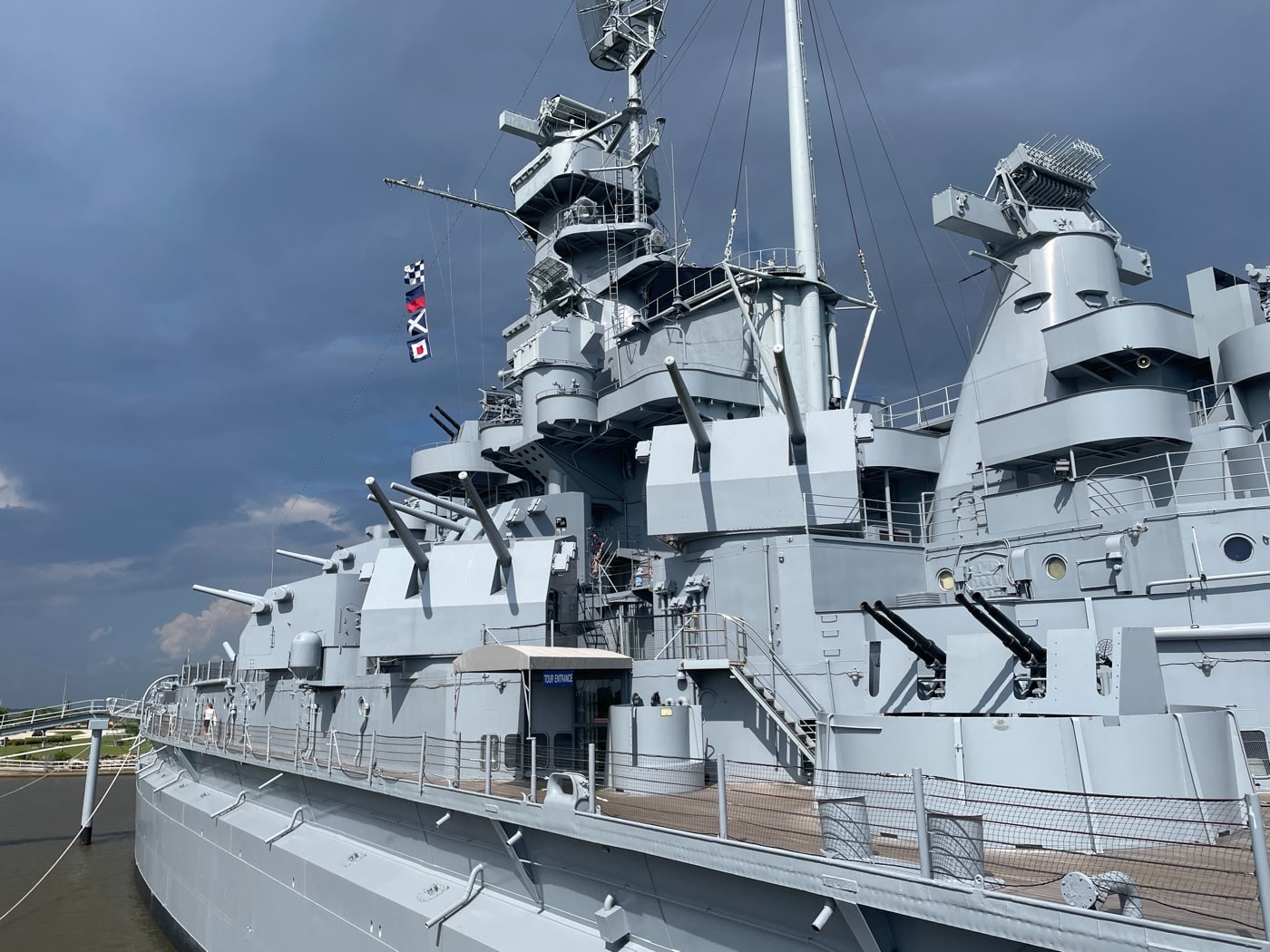 Image of the USS Alabama’s deck showing several of her ten twin 5-inch gun turrets arranged along the sides of the ship. The photo captures the clean lines and sturdy construction of the mounts, with the barrels pointed outward or at varying angles for training. Background details include other weapons, superstructure elements, and possibly the ocean or dock if taken at the memorial. Visitors may be present for scale, but the focus is on the turrets themselves. The lighting highlights the texture and size of the gun mounts. This photo provides context for how these weapons were positioned for all-around defense. It’s a clear demonstration of the Alabama’s layered firepower.