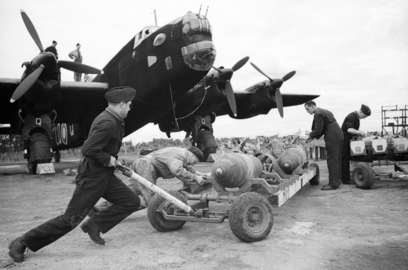 British ground crew load 1,000-pound bombs into the bomb bay of a Handley Page Halifax heavy bomber during World War II. Several men work with bomb trolleys and lifting equipment to carefully maneuver the large bombs beneath the aircraft. The Halifax’s long bomb bay allowed it to carry multiple heavy weapons for strategic raids. The scene shows the scale of the bombs compared to the crew, underlining the destructive power the Halifax could deliver. The aircraft is stationary on an airfield with its undercarriage visible as the loading process takes place. This preparation was a routine but crucial part of RAF operations. The image captures the teamwork and precision required on the ground before each mission.