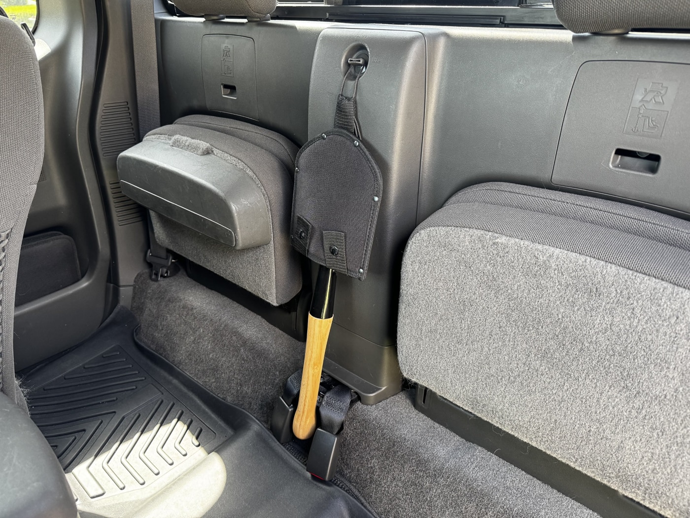 The Cold Steel Special Forces Shovel secured inside a pickup truck, positioned between seats for quick access. The black nylon sheath covers the sharpened blade, with two snap closures keeping it in place. A loop of paracord is attached to the sheath’s webbing strap, allowing it to hang from a latch or hook in the truck cab. The hickory wood handle is visible extending from the sheath, showing the tool’s compact size. The image illustrates how the shovel can be kept as part of a vehicle emergency kit. Its placement makes it easy to retrieve in off-road or roadside situations.