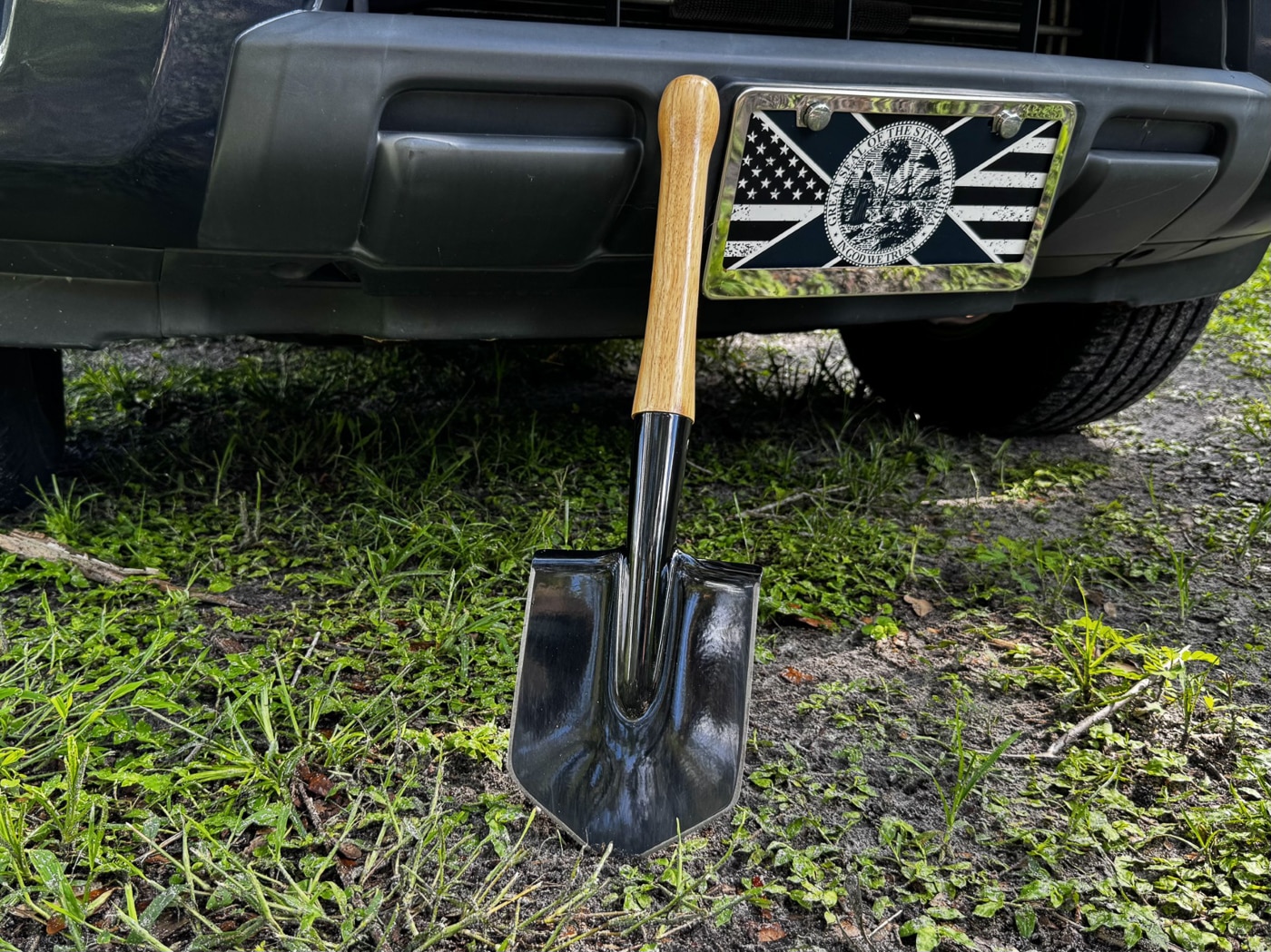 The Cold Steel Special Forces Shovel leaning upright against the side panel of a pickup truck. The shovel has a coated carbon steel blade with sharpened edges on all three sides. Its hickory wood handle is fixed securely into the socket with two large screws. The blade’s compact, slightly concave shape mirrors the Soviet MPL-50 design. The truck provides scale, showing that the shovel’s overall length is around 20 inches, making it portable for transport. The image suggests the tool’s role as part of a vehicle emergency kit or for use during off-road trips.