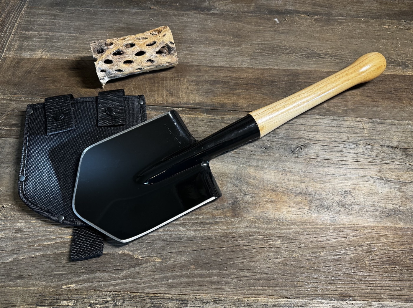 A Cold Steel Special Forces Shovel resting next to its fitted nylon sheath, photographed on a flat surface. The shovel’s carbon steel blade is clearly visible, with sharpened edges along both sides and the top. The hickory wood handle is secured into the socket with two large Phillips screws. The sheath has two retention snaps to hold the blade and a webbing strap for mounting. The metal blade is painted for corrosion resistance, but the edge grind is exposed, showing its readiness for chopping. The compact size makes it suitable for portability in vehicles or packs.
