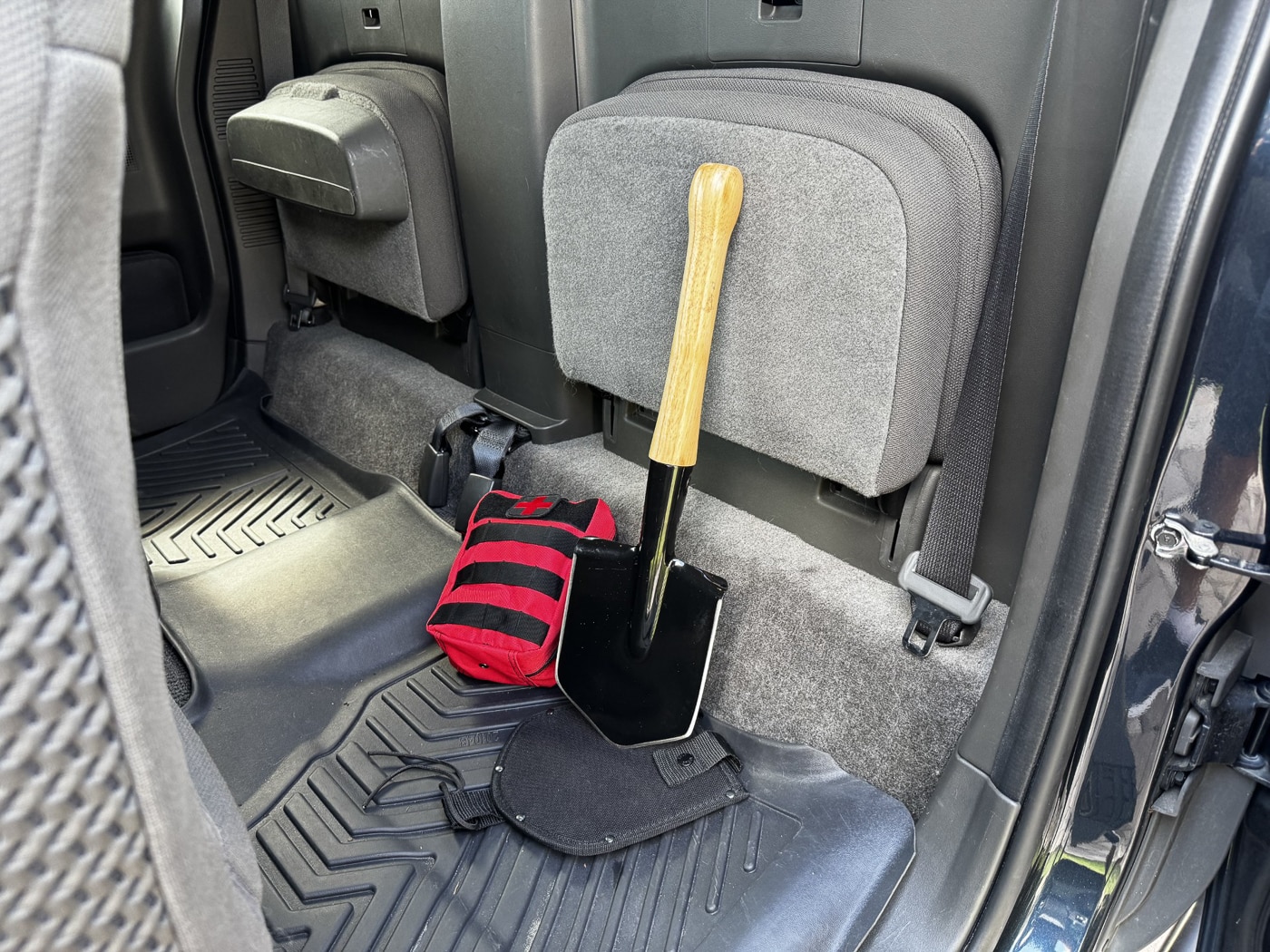 The Cold Steel Special Forces Shovel positioned next to an Individual First Aid Kit inside a vehicle interior. The shovel is in its black nylon sheath with retention snaps, while the IFAK is in a compact zippered pouch. Both are stored within reach, suggesting quick deployment in an emergency. The shovel’s hickory handle extends from the sheath, and the mounting loop is attached to a fixed point in the cab. The IFAK is secured in place to prevent movement during travel. The arrangement reflects a balanced approach to vehicle preparedness, combining a versatile tool with essential medical supplies.