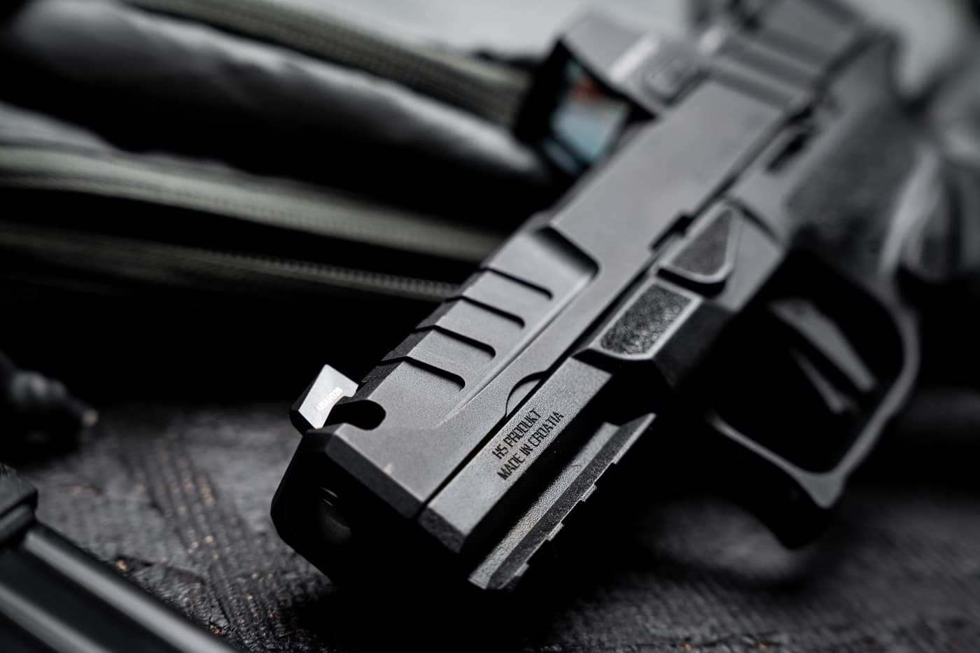 A close-up photograph of the Springfield Armory Echelon 4.0C Comp’s billet-machined slide, focusing on the integral single-port compensator cut. The port is positioned at the slide’s front, directly behind the tritium/luminescent front sight, and is precisely aligned with the barrel’s matching cut. The matte black Melonite finish highlights the sharp machining lines and clean edges around the opening. This design allows high-pressure gases to vent upward, countering muzzle rise during firing. Light catches on the surface, showing the depth and shape of the cut. The image emphasizes the factory-engineered quality and functional purpose of the compensator in improving shooting control.