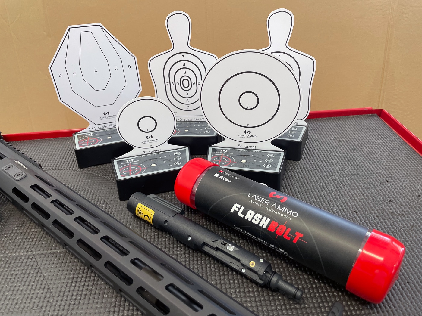 The Laser Ammo Flash Bolt training device and several i-MTTS interactive targets arranged on a tabletop before being set up. The Flash Bolt is a bolt carrier group replacement for AR-15 rifles, shown alongside its trigger activation switch and connecting cable. The i-MTTS targets are mounted on their black electronic bases with reflective faces in different shapes and sizes. This layout shows the system before deployment in a home or range environment. The combination allows safe dry-fire training with visual and audio hit confirmation. The components are organized and ready for placement in a training area.