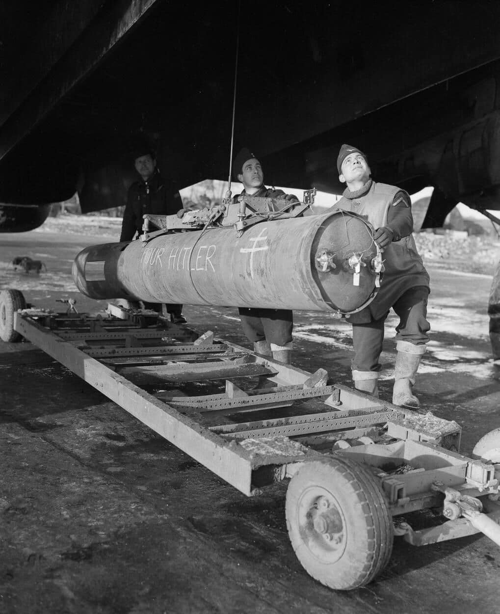 Free French ground crews load a 2,000-pound high-capacity bomb into the bomb bay of a Handley Page Halifax bomber during World War II. Several men work together to maneuver the large bomb into place beneath the heavy aircraft. The Halifax’s spacious bomb bay allowed it to carry weapons of this size alongside smaller bombs. The presence of Free French crews demonstrates the cooperation between Allied forces in preparing and flying these missions. Heavy ordnance such as the 2,000-pound bomb was typically employed against hardened or strategic targets. The Halifax looms overhead with its landing gear down, showing the scale of both the aircraft and the bomb being loaded. This image illustrates the logistical effort required to keep bombers armed and operational.
