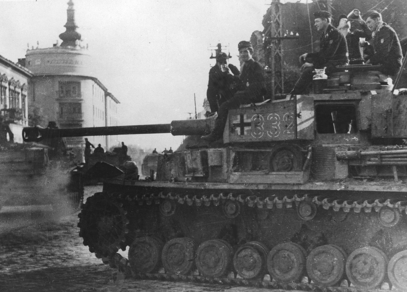 The photograph shows a German Panzer IV Ausf. H tank parked or moving slowly along a cobblestone street in Debrecen, Hungary, during World War II. The tank features full Schürzen side armor skirts along the hull and turret, as well as the long-barreled 75mm KwK 40 L/48 gun with a muzzle brake. Buildings with European architectural style line both sides of the street, some with shuttered windows and signs of wartime wear. The road is relatively narrow, emphasizing the size of the vehicle in the urban environment. Lighting suggests daytime conditions, with shadows cast to one side. The image conveys the presence of heavy armor within a civilian cityscape during active operations on the Eastern Front.