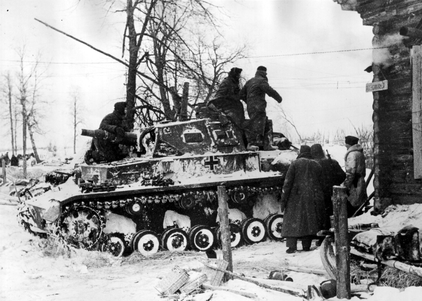 The photograph shows a German Panzerkampfwagen IV Ausf. E tank positioned next to a wooden house in a rural area on the Eastern Front during World War II. The vehicle features the short-barreled 75mm KwK 37 main gun and relatively thin armor typical of early-war Panzer IV models. The tank’s boxy hull and large road wheels are clearly visible, and no additional side armor skirts are present. The wooden structure beside it has a simple pitched roof and plank walls, typical of rural Soviet architecture. The surrounding ground is unpaved, with sparse grass and dirt. This image captures the contrast between military hardware and civilian structures in war-torn Eastern Europe.