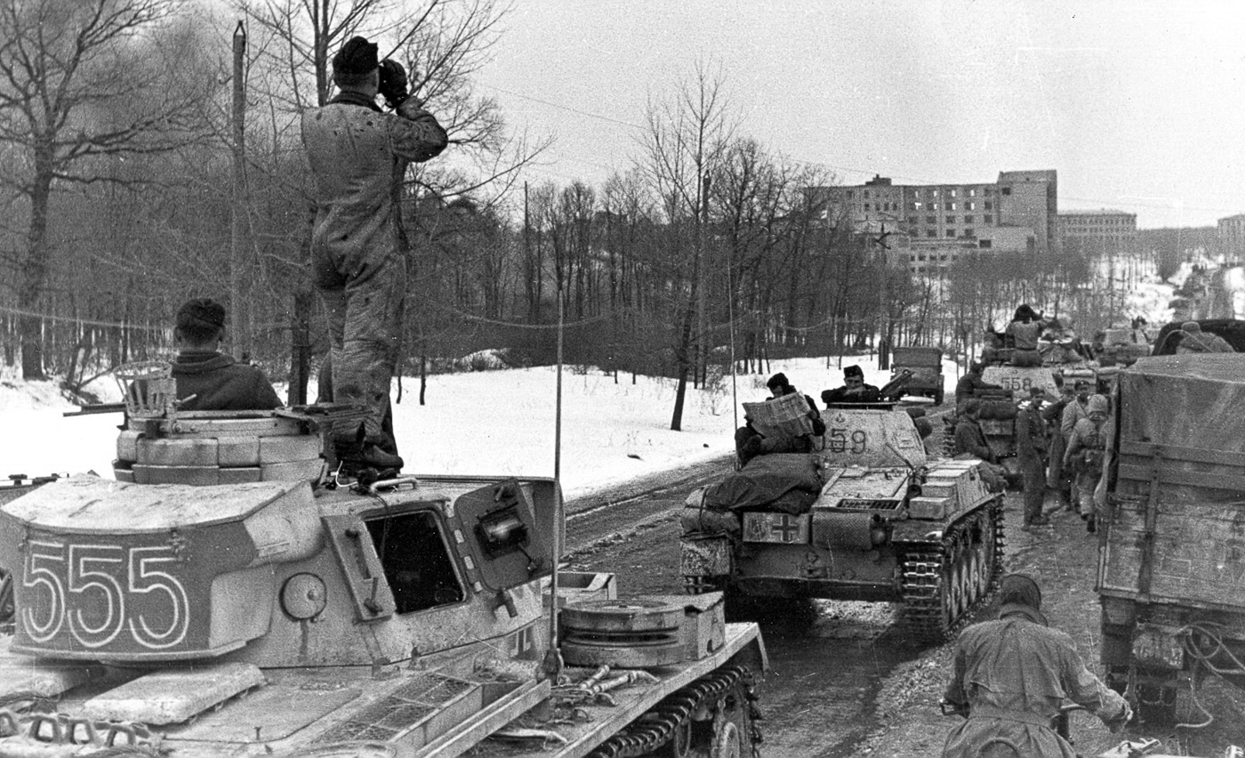 The photograph shows a German tanker standing on the turret of a Panzer IV tank near Kharkiv, Ukraine, during World War II. The soldier is dressed in standard black Panzer crew uniform and holds a pair of binoculars to his eyes, scanning the distance for enemy movement. The Panzer IV appears to be a mid-to-late-war variant with a long-barreled 75mm KwK 40 L/48 gun and Schürzen side armor skirts. The surrounding terrain is open, with low grass and a distant treeline visible under a clear sky. The tanker’s elevated position provides a wide field of view, critical for spotting Soviet troops in the area. The image conveys both the tactical awareness and vulnerability of tank crews operating in open steppe environments.