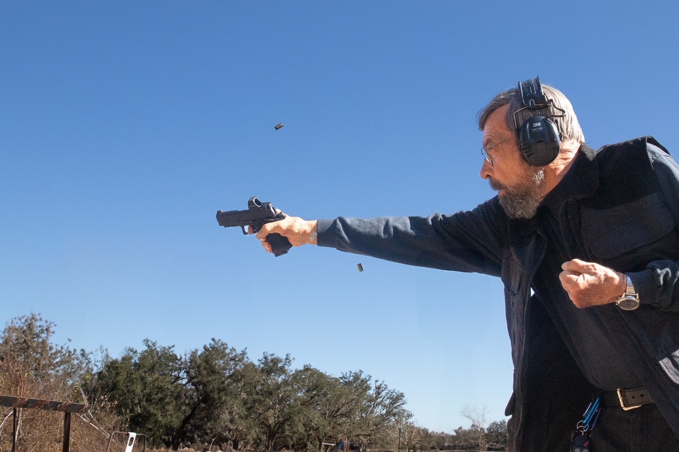 This image shows Massad Ayoob firing the Springfield Armory Prodigy DS 1911 rapidly with one hand. The pistol is extended fully as brass is seen ejecting, indicating live fire in progress. The Prodigy is a double-stack 1911 designed to offer higher capacity than the traditional single-stack configuration. It retains the recognizable ergonomics of the 1911 platform while incorporating modern features like an optics-ready slide and aggressive grip texturing. Ayoob’s one-handed shooting demonstration emphasizes the pistol’s controllability even during rapid fire. The image captures both recoil management and the speed of follow-up shots. The background reflects an outdoor shooting range environment, reinforcing the training and live-fire context.