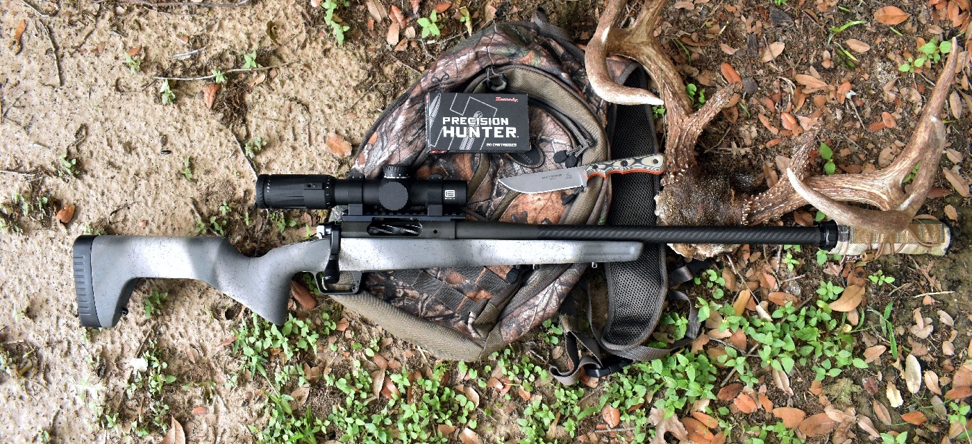 The photograph shows a TOPS Muley Skinner knife placed next to an unloaded Springfield Armory Redline rifle and a hunting backpack, all resting on the ground in an outdoor setting. The equipment appears ready for a deer hunt, with the knife’s orange-lined G10 handle clearly visible for contrast against the earth-toned backdrop. The rifle is laid parallel to the pack, with the knife positioned for easy access. This composition highlights a realistic collection of gear used by hunters, as referenced in the TOPS Muley Skinner knife review. The image is designed to give context to the article’s discussion of reliable hunting tools. Background details include leaves, dirt, and possibly some grass, all emphasizing a typical field scenario.