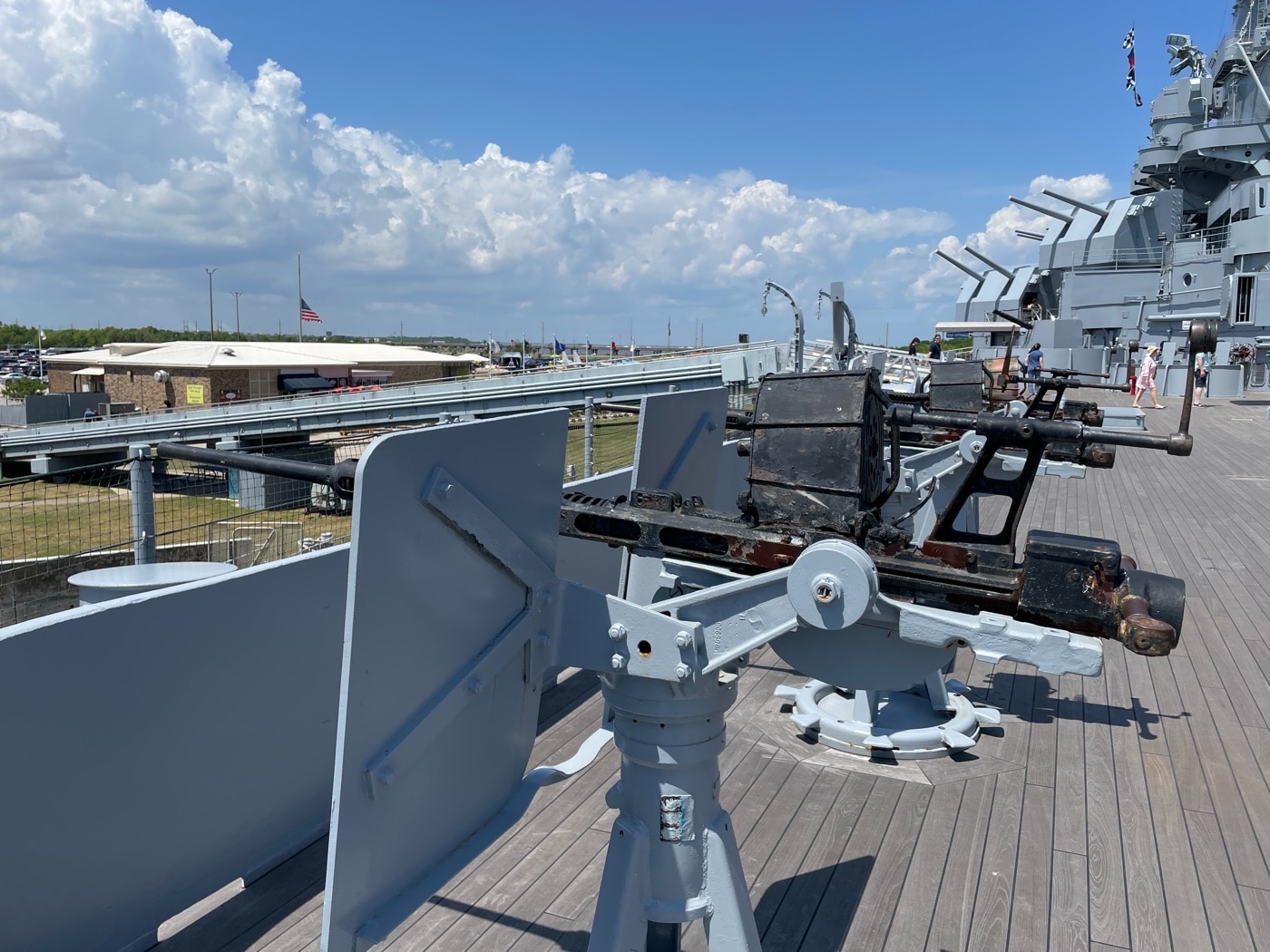 Photo showing a single Oerlikon 20mm antiaircraft cannon mounted on the deck or superstructure of USS Alabama. The weapon features a slim barrel, open breech, and distinctive drum magazine mounted on top. Hand grips, aiming sights, and a shoulder stock are visible, emphasizing the gun’s design for rapid, direct fire. The mount is exposed, allowing for a wide range of motion to track fast-moving targets. Background details include metal railings, deck plating, and possibly the sea or other superstructure elements. No crew is present, keeping the focus on the gun itself. The image highlights the Alabama’s extensive use of these small, fast-firing weapons for close-in air defense. This scene illustrates the practical layout and technical features of World War II naval antiaircraft guns.