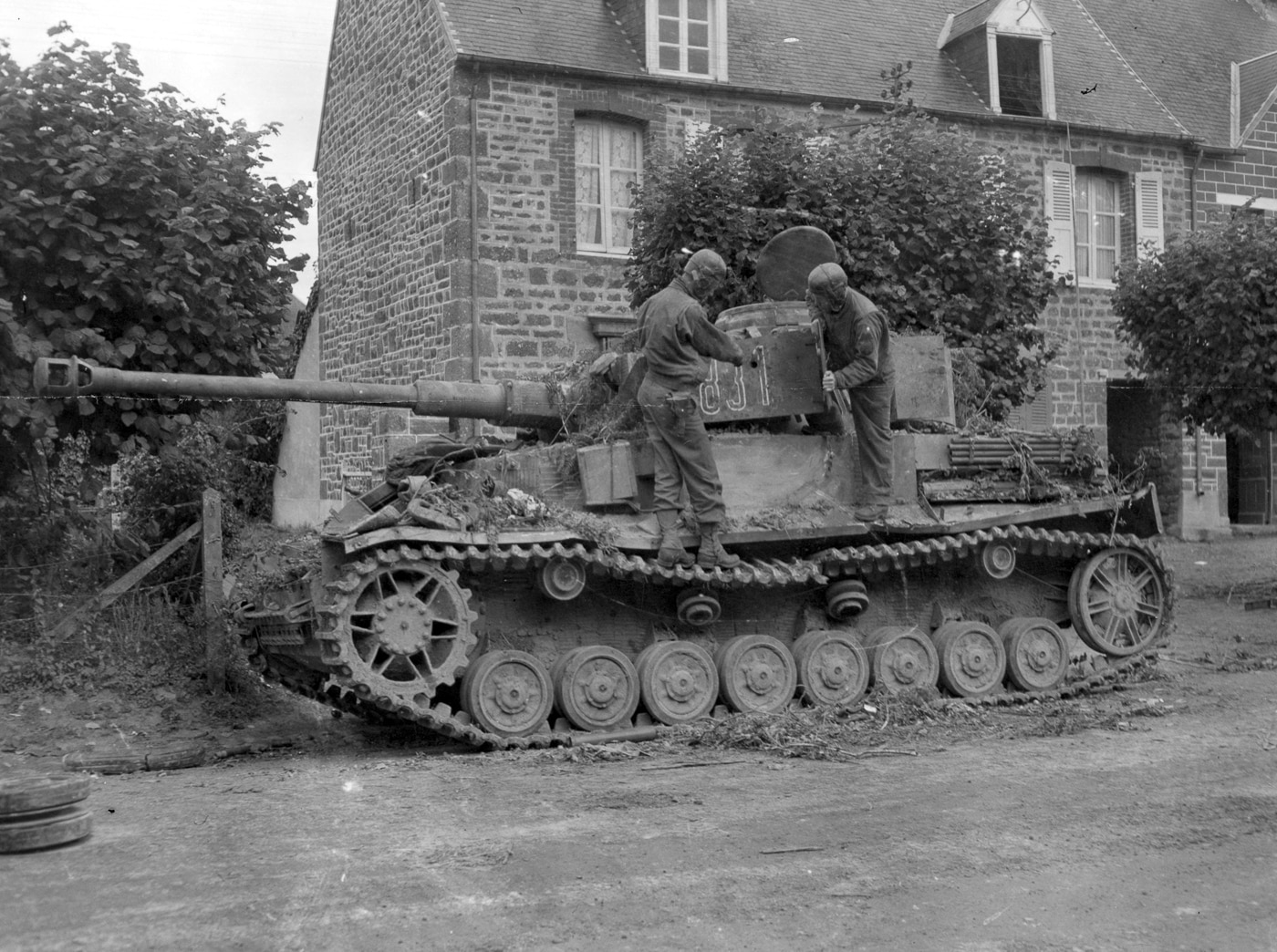 The image shows a destroyed German Panzer IV tank positioned on a narrow street in Saint-Denis-le-Gast, France, during World War II. The tank has sustained visible damage from a 37mm artillery shell, which disabled it during combat. Its tracks remain intact, but the front armor shows an impact mark, and the gun barrel is angled slightly downward. Surrounding buildings exhibit traditional French village architecture, with stone walls and shuttered windows. Debris is scattered near the vehicle, and no crew are present. The scene captures the aftermath of a tank being neutralized in close-quarters urban fighting during the Normandy campaign.