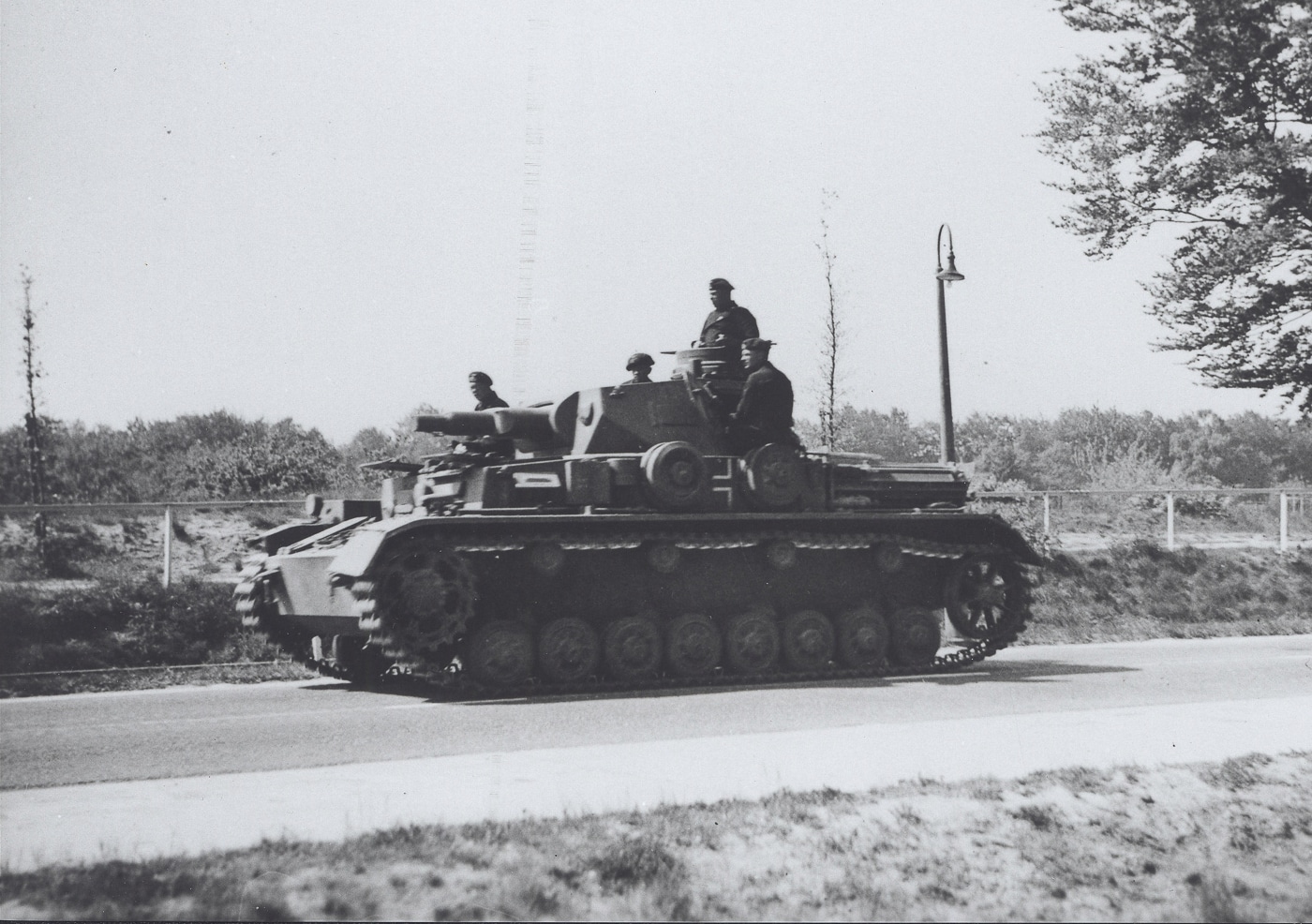 The photograph shows a German Panzer IV tank driving down Sint Annastraat during the German invasion of the Netherlands in May 1940. The tank, an early-war variant, is equipped with a short-barreled 75mm KwK 37 gun and minimal side armor. It moves along a paved urban street lined with multi-story brick buildings, some showing signs of damage. Civilians can be seen at a distance, either watching or retreating from the approaching vehicle. The tank’s tracks are kicking up small amounts of dust as it advances. This street-level view captures the shock and immediacy of armored vehicles entering a city during the campaign.