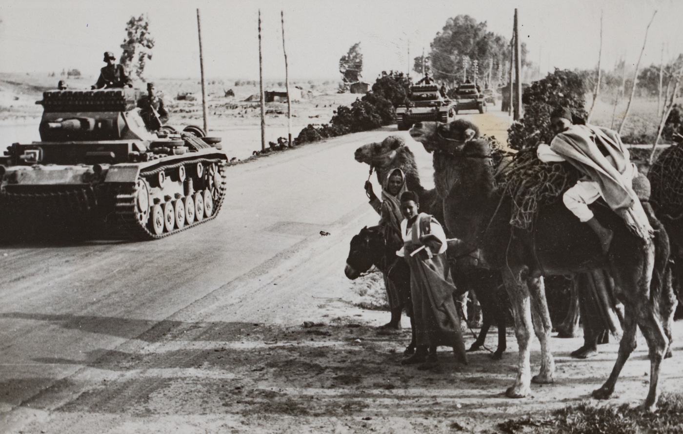 The photograph shows several German Panzer IV tanks from the Afrika Korps advancing along a dusty road in Libya during World War II. The tanks, painted in desert camouflage, are equipped with short-barreled 75mm KwK 37 guns typical of early-war models used in North Africa. Along the roadside, local civilians and riders on camels watch the column pass, some standing while others remain mounted. The background reveals open desert terrain with a clear sky. The juxtaposition of heavily armored vehicles and traditional desert transport animals emphasizes the cultural and technological contrasts of the North African campaign.