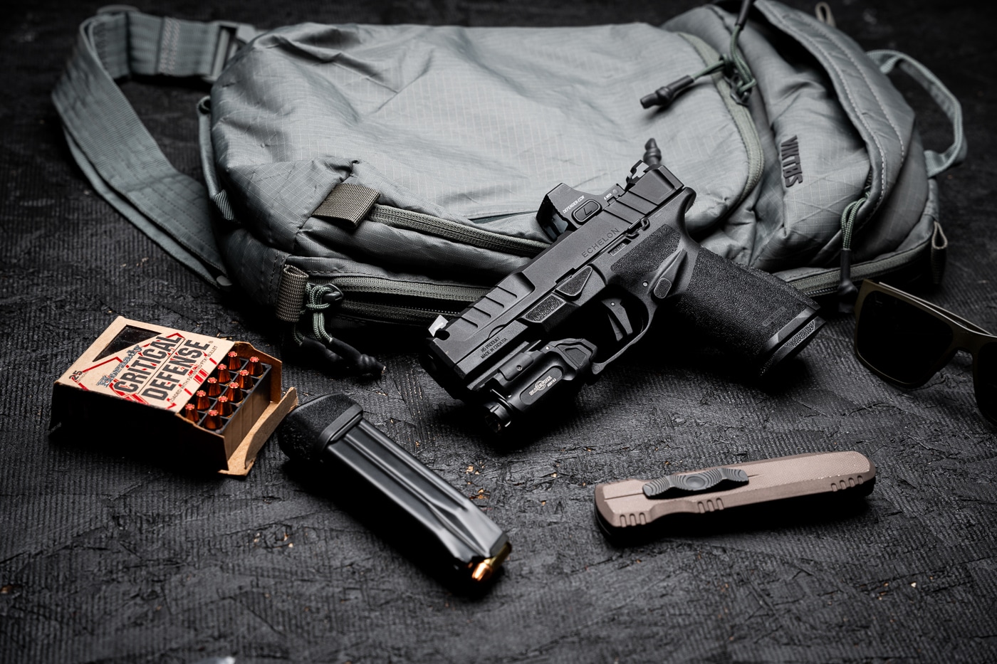 A gear layout featuring the Springfield Armory Echelon 4.0C Comp 9mm pistol positioned next to a backpack and several boxes of 9mm ammunition. The pistol has a black Melonite-finished slide with an integral single-port compensator, a 4-inch hammer-forged barrel, and a mounted red dot optic co-witnessed with tritium U-Dot sights. The Adaptive Grip Texture on the frame is clearly visible, designed for secure handling. The backpack is placed to suggest discreet transport or off-body carry. The ammunition boxes are arranged nearby, indicating readiness for range sessions or defensive use. The overall composition emphasizes the Echelon 4.0C Comp as part of a practical, portable shooting setup that combines performance, concealability, and preparedness.