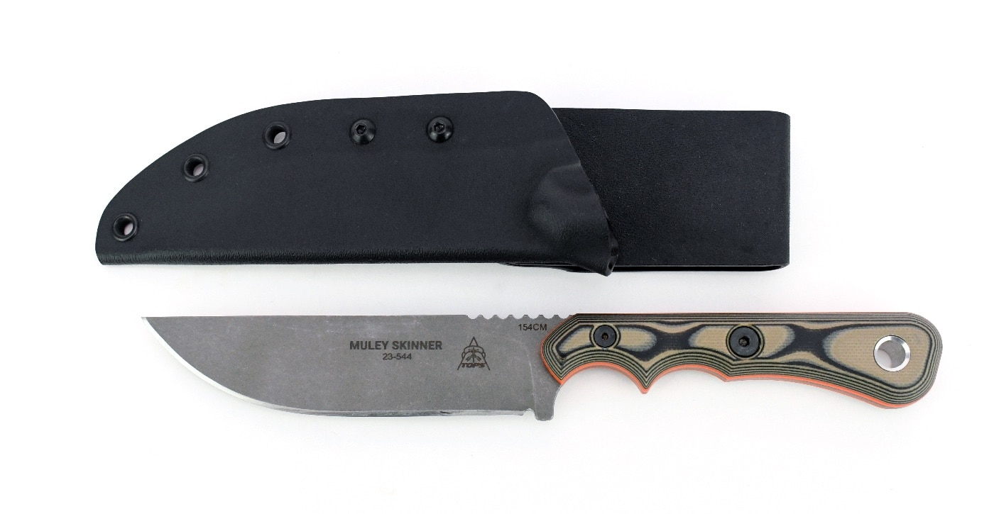 The image displays the TOPS Muley Skinner knife positioned next to its black kydex sheath on a neutral surface. The knife’s 4.5-inch curved blade and black-and-tan G10 handle with bright orange liners are clearly visible, along with the lanyard ring and Torx screws in the grip. The kydex sheath is molded specifically to fit the knife, providing maximum retention and preventing accidental slips — a key advantage mentioned in the TOPS Muley Skinner knife review. The sheath features rivets or eyelets for attachment to belts or gear. This picture highlights the practical carry solution for hunters and outdoorsmen. Both the knife and sheath are shown clean and ready for use. The overall setup is compact and field-ready, aligning with the review’s emphasis on reliability and safety.