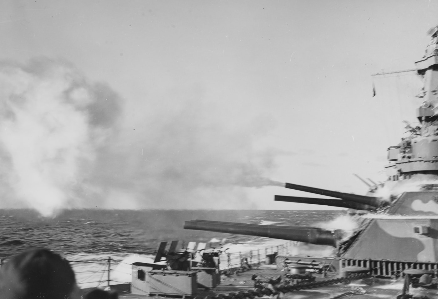 Color or black and white photo showing the 16-inch guns of the USS Alabama (BB-60) firing during a live-fire exercise or combat operation. The image captures the moment the guns discharge, with massive bursts of flame and thick smoke erupting from the barrels. The shockwave distorts the air around the muzzle, while the recoil pushes the gun barrels slightly backward. Shell casings and debris can be seen on the deck below the turrets. The rest of the ship’s superstructure is partially visible, but the main focus is on the enormous firepower on display. This image demonstrates the destructive capability and dramatic visual impact of the battleship’s main battery. The scene highlights both the engineering and combat power of World War II-era naval artillery.