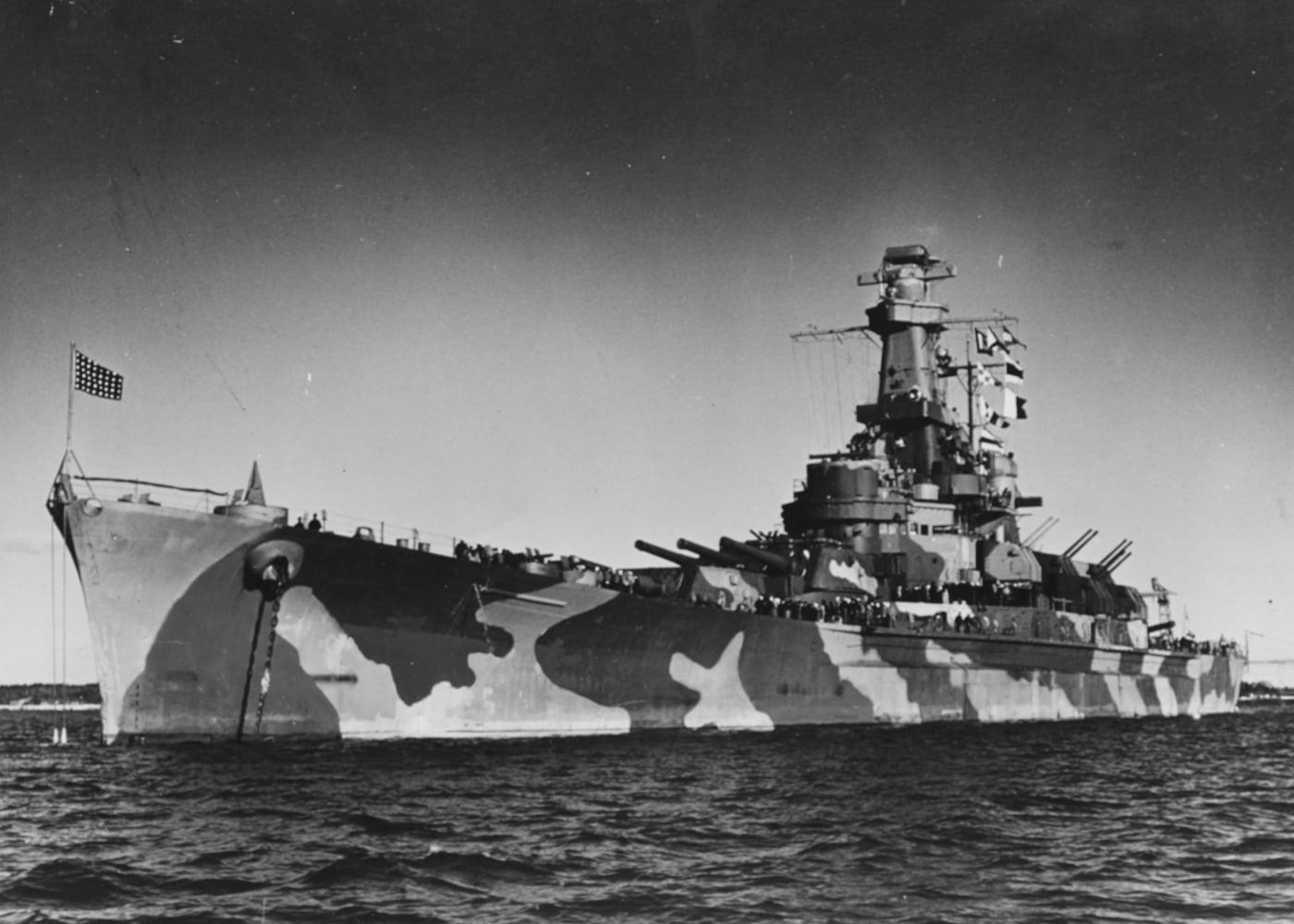 Historic black and white photo of the USS Alabama (BB-60) anchored in Casco Bay, Maine, in 1942, shortly after her commissioning. The battleship is seen in profile, with her main gun turrets facing forward and secondary armament visible along the superstructure. Calm water reflects the ship’s hull and superstructure, with land and trees faintly visible in the background. Smoke or steam may be rising from the ship’s stacks, indicating operational readiness. The scene conveys a sense of quiet before active deployment. The overall composition emphasizes the scale of the ship in relation to the peaceful bay. This image documents the Alabama’s early days as a newly completed battleship preparing for wartime service.