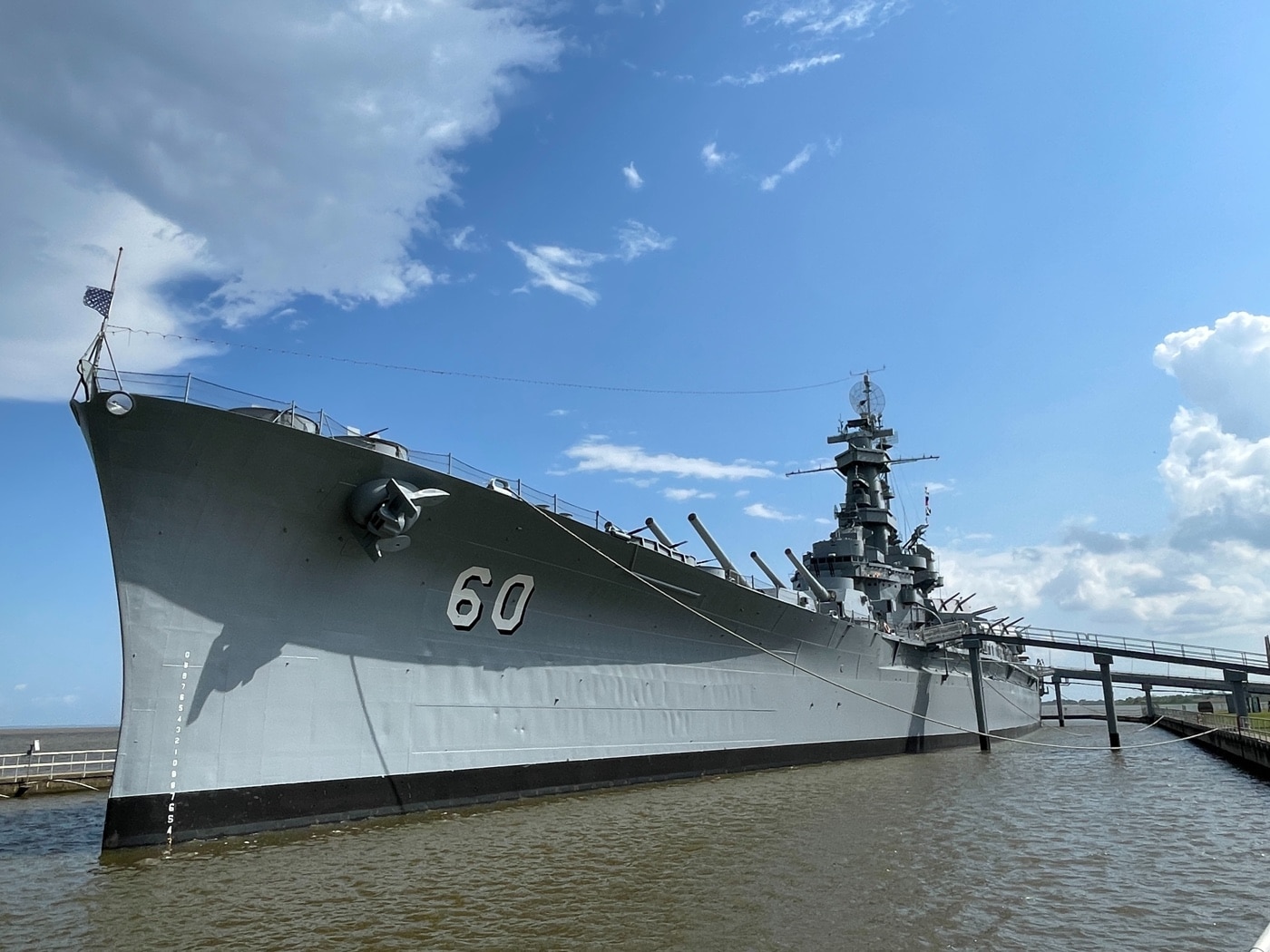 Color photo taken at the USS Alabama Memorial Park showing the battleship docked along the waterfront. The image highlights the enormous size of the ship, with the hull stretching far into the distance and towering above visitors on the walkway below. The ship’s gray steel plating, tall superstructure, and numerous gun turrets dominate the frame. People on the pier appear tiny in comparison, emphasizing the ship’s scale. Flags may be flying along the deck, and informational signs or displays can be seen nearby. The sky is bright and clear, adding to the dramatic effect. This scene gives a real sense of the Alabama’s massive presence as a preserved World War II warship and historical landmark.