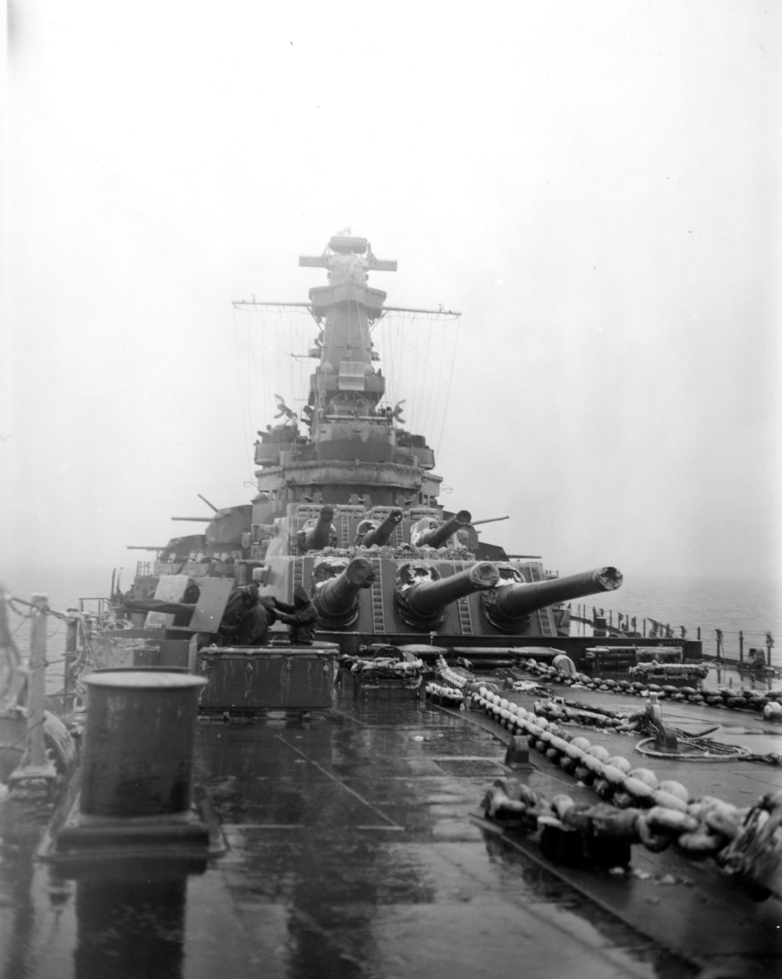 Black and white photo taken from the bow of USS Alabama (BB-60) during a snowstorm on her shakedown cruise in January 1943. The camera looks aft along the ship’s deck, with three 16-inch main gun turrets visible, all pointing toward the rear. Snow has accumulated heavily on the deck and turret roofs, creating a stark contrast against the dark metal of the ship. The harsh weather obscures some details, adding to the sense of cold and adversity. The main battery’s sheer size dominates the foreground, emphasizing the ship’s firepower even in adverse conditions. Snowfall reduces visibility, making the image feel both harsh and quiet. This scene illustrates the challenges faced by the crew as the battleship was tested in winter conditions.