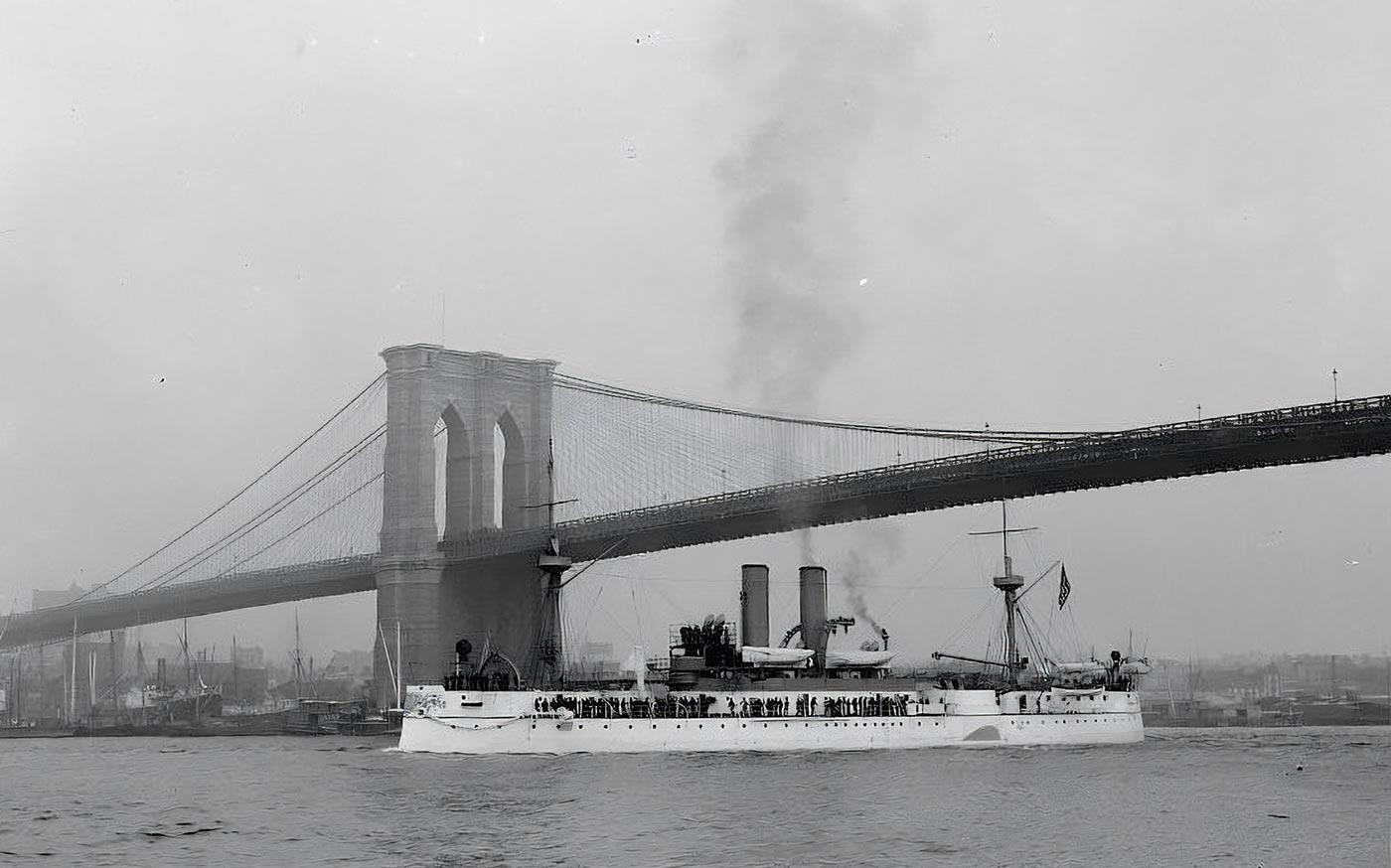 A historic photograph of USS Maine passing beneath the Brooklyn Bridge in New York City. The warship’s armored hull, gun turrets, and tall masts are visible as she moves along the East River. Above, the massive suspension bridge dominates the skyline, representing one of the era’s great engineering achievements. The image juxtaposes the Navy’s new steel battleship with a modern urban landmark. Crowds can be faintly seen on the bridge, possibly watching the vessel’s passage. The picture reflects both the pride in America’s growing naval power and its industrial advancement. It remains one of the most iconic photographs of USS Maine before her destruction.