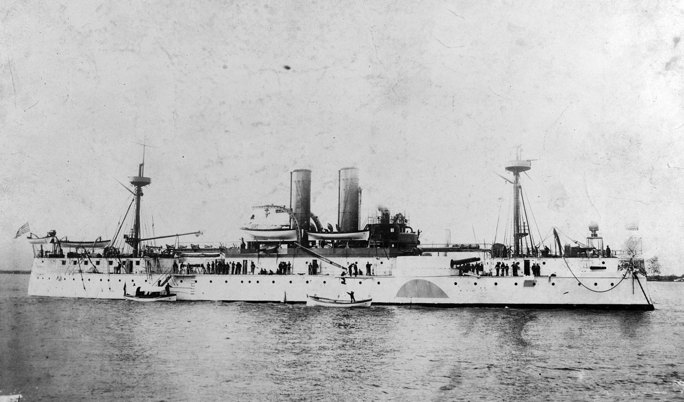 A historic photograph showing the USS Maine from the starboard side while afloat. The battleship’s armored hull stretches across the water, with its forward and aft gun turrets visible along the deck. The ship’s twin masts rise above the superstructure, a reminder of the transitional design between sail and steam power. The photo captures the Maine at rest, before her fatal deployment to Havana Harbor. Details of the ship’s secondary armament and smokestacks can be seen clearly from this angle. This image highlights the naval architecture of the late 19th century. It remains one of the most recognized photos of the vessel prior to its destruction.