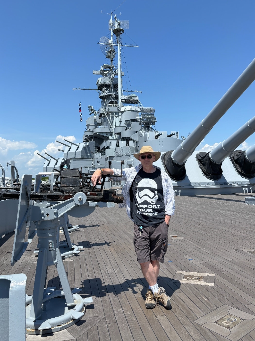 Color photo showing the author standing on the open deck of the USS Alabama, docked at her memorial park in Mobile, Alabama. The author is in casual clothing, possibly wearing a hat or sunglasses, with the battleship’s superstructure, gun turrets, or deck equipment visible in the background. The weather appears clear and sunny, with blue sky and bright light reflecting off the ship’s steel surfaces. Other visitors may be present at a distance, but the author is the main focus of the shot. Details such as railings, hatches, or museum signage add context to the historic setting. This image highlights the opportunity for guests to physically explore the restored World War II battleship. The scene helps personalize the visitor experience aboard the Alabama.
