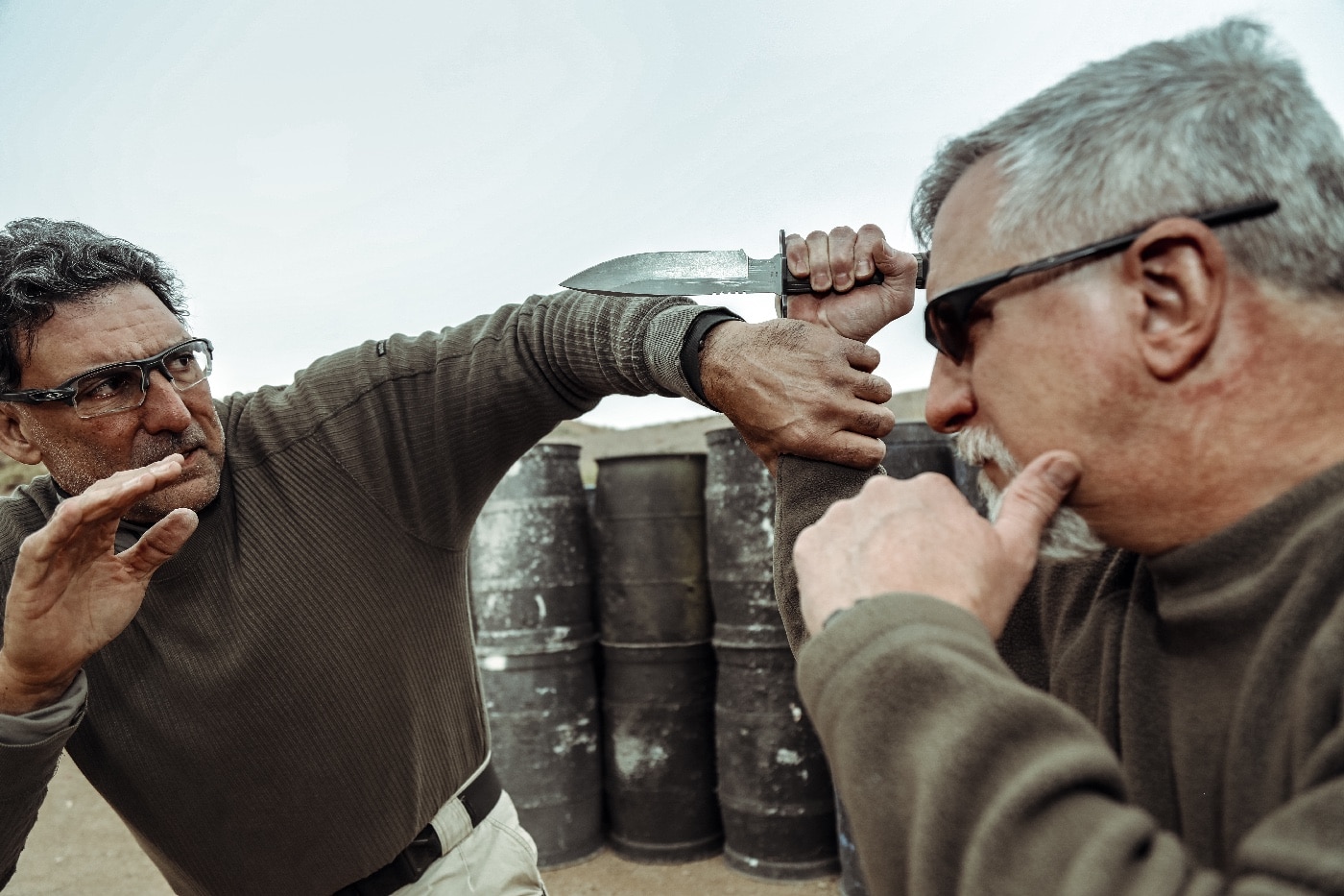 The author is pictured mid-action, physically defending himself against an attacker armed with a knife. His body is angled defensively, one hand intercepting the knife arm while the other prepares to counter or disarm. Both individuals are focused and intense, their faces and body language showing the urgency of the situation. The background is a neutral training space, with minimal distractions to keep attention on the defense technique. This image illustrates practical self-defense against edged weapon threats, highlighting the need for rapid reaction and hard skills training to overcome the power curve in a violent confrontation. It’s a realistic depiction of close-quarters combat, emphasizing the value of repetition and professional instruction. Perfect for articles on knife defense, reaction time, and personal protection.