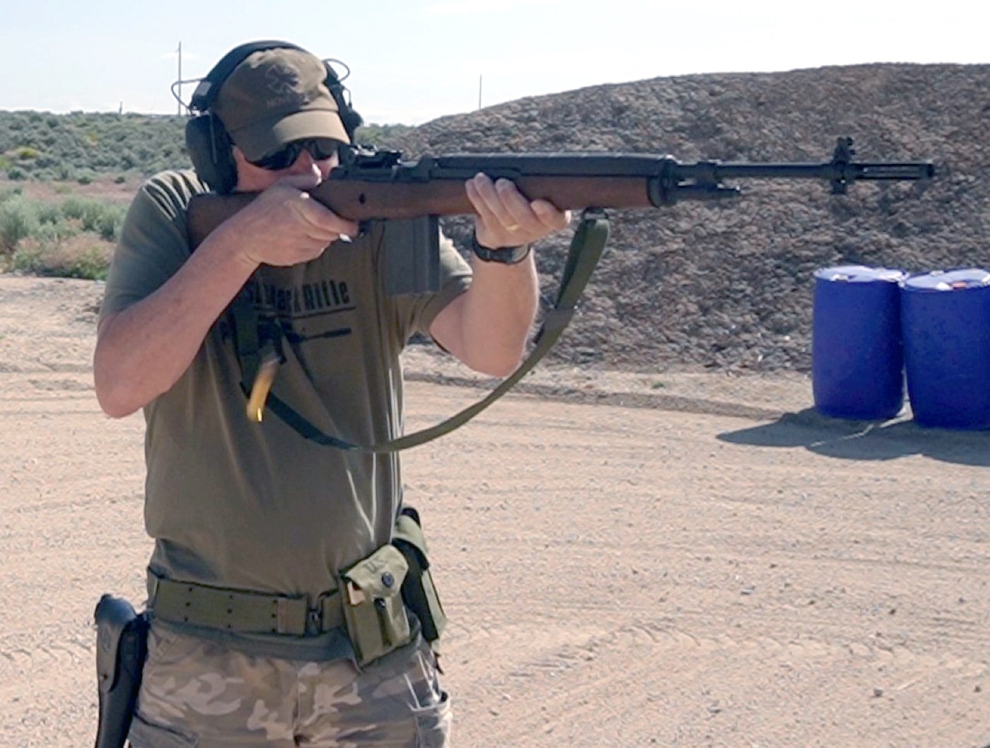 The image shows the author shooting the Springfield Armory M1A rifle at an outdoor shooting range, standing behind the firing line in a typical shooting stance. The author is aiming down the iron sights, with the rifle’s walnut stock pressed against the shoulder. Spent shell casings may be seen ejecting from the action, indicating live fire. Range equipment, such as benches, targets, and berms, appear in the background, along with standard outdoor safety features. The author is wearing appropriate eye and ear protection for safety. The photo provides a clear view of the M1A’s size, wood furniture, and overall layout while in use. This image works well for articles discussing the M1A’s handling, range performance, or suitability for collectors and shooters interested in U.S. military-style rifles.