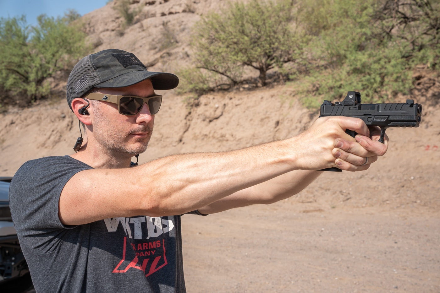 The author is captured mid-shot while firing the Springfield Armory Echelon 4.0C Comp 9mm pistol at an outdoor shooting range. The compact handgun has a black Melonite-finished slide with an integral single-port compensator machined directly into the barrel and slide nose. A mounted red dot optic is visible, paired with factory tritium U-Dot sights. The shooter maintains a two-handed grip, showcasing the Adaptive Grip Texture for secure handling. The background features range berms and natural lighting typical of an outdoor range. Spent brass is in motion, highlighting the pistol’s ejection cycle. The scene emphasizes the Echelon 4.0C Comp’s blend of concealability, recoil control, and performance-focused design.
