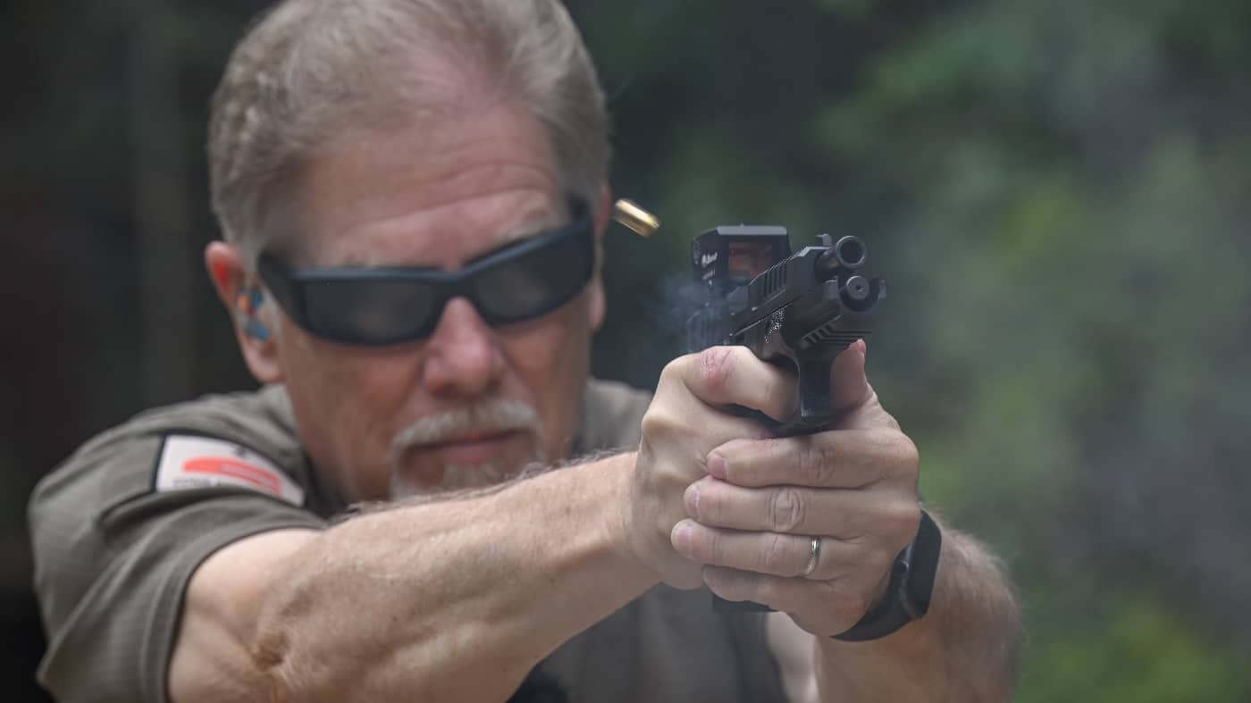 Photo shows a Springfield Armory Echelon pistol mid-recoil as it is fired by the author, with a Burris FastFire E enclosed red dot sight mounted on the slide. The slide is visibly cycling to the rear, and a spent casing is ejecting, illustrating the moment of recoil. The FastFire E’s robust, enclosed housing remains securely attached despite the sharp motion, highlighting its durability under stress. The author’s hands maintain a solid grip on the pistol, and the background features a typical range setting with targets and barriers. The optic’s lens and controls are clearly visible, showing no signs of shifting or damage from repeated firing. Lighting and angle keep focus on both the optic and the dynamics of recoil. This photo supports the review’s emphasis on the FastFire E’s toughness and reliability under real shooting conditions.
