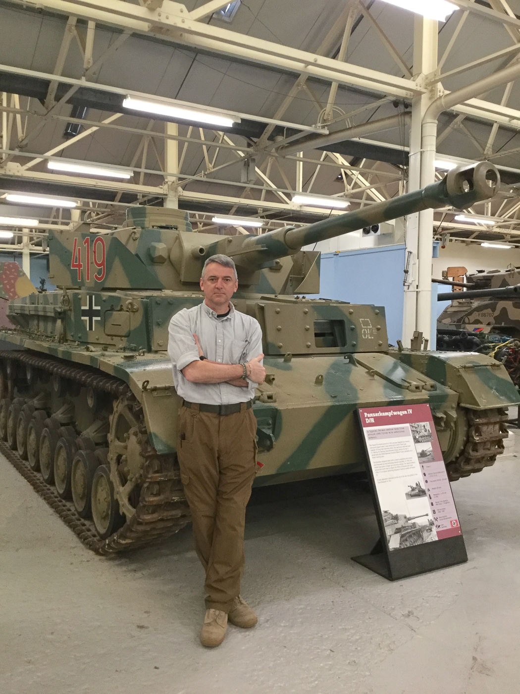 The photograph shows the author, Will Dabbs, standing beside a Panzer IV Ausf H medium tank displayed at an outdoor section of a military museum. The tank retains its Schürzen side armor skirts and the long-barreled 75mm KwK 40 L/48 gun with a prominent muzzle brake. Its dark paint scheme appears well-preserved, suggesting restoration. The author, dressed in casual clothing, stands near the left track, giving a clear sense of the tank’s size and height. The background includes museum signage and a paved walkway, indicating a public exhibit space. Lighting conditions are bright, casting shadows that enhance the tank’s structural details.